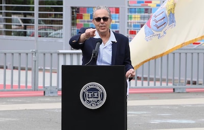 A man wearing sunglasses and a suit jacket points forward while standing at a podium with a colorful building and a flag in the background.