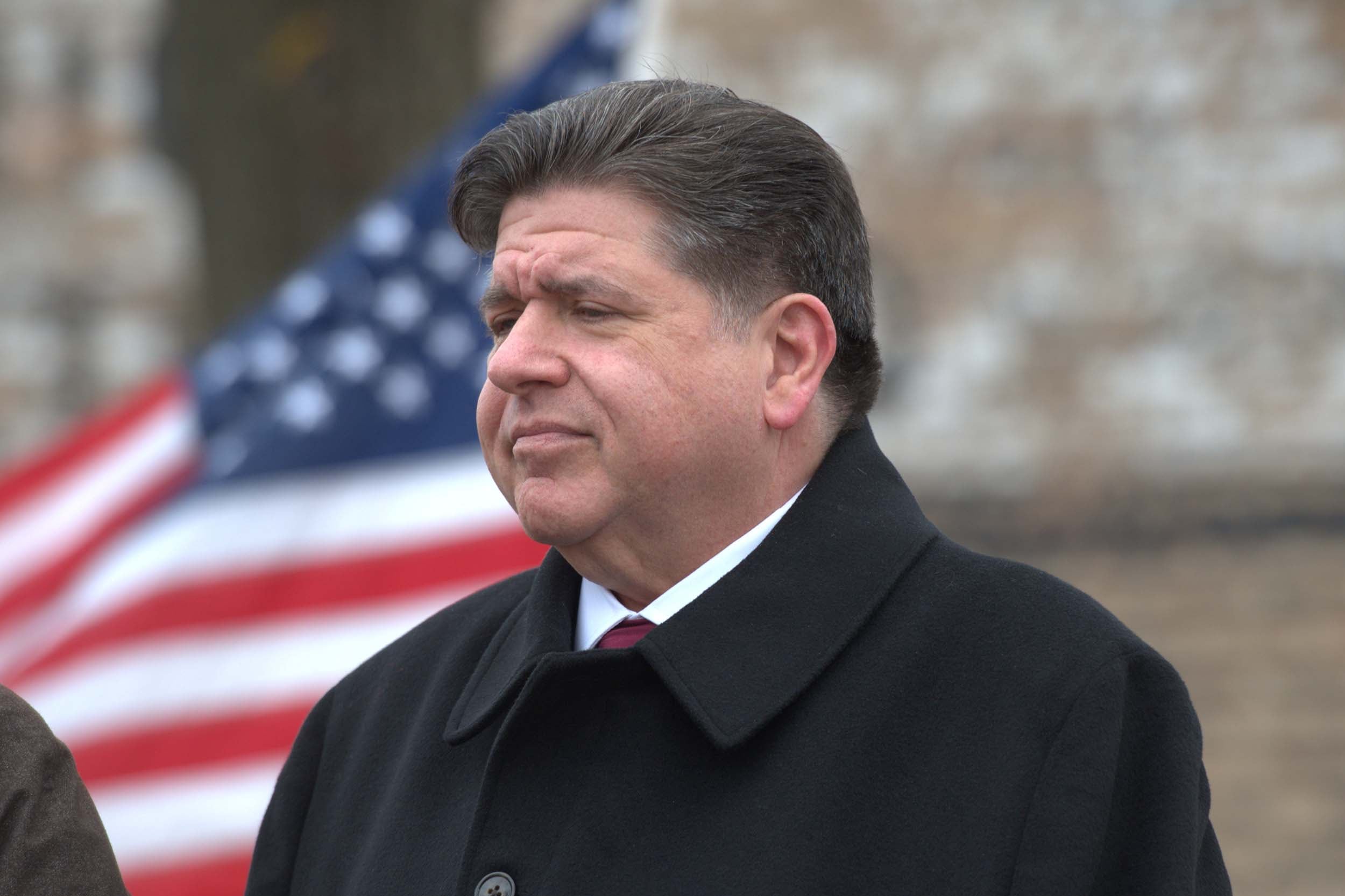 A photograph of a white man in a suit and black jacket standing outside in front of an American flag.