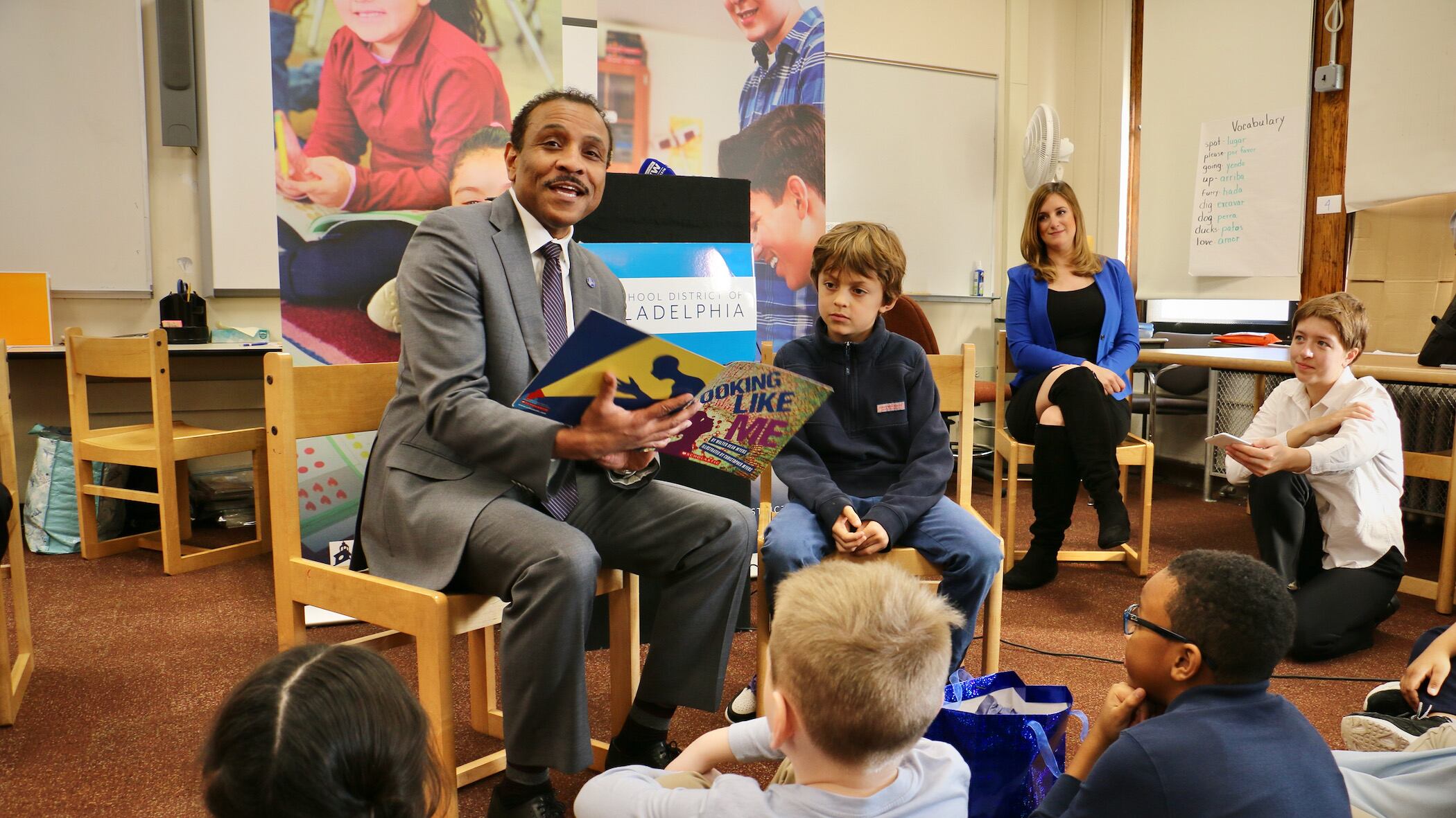 A man in a gray suit and dark tie sits on a chair reading a book aloud. A boy in a blue shirt and jeans sits next to him. Students sit on the floor listening. A teacher wearing a black dress, black boots, and a bright blue jacket sits to the left of him.