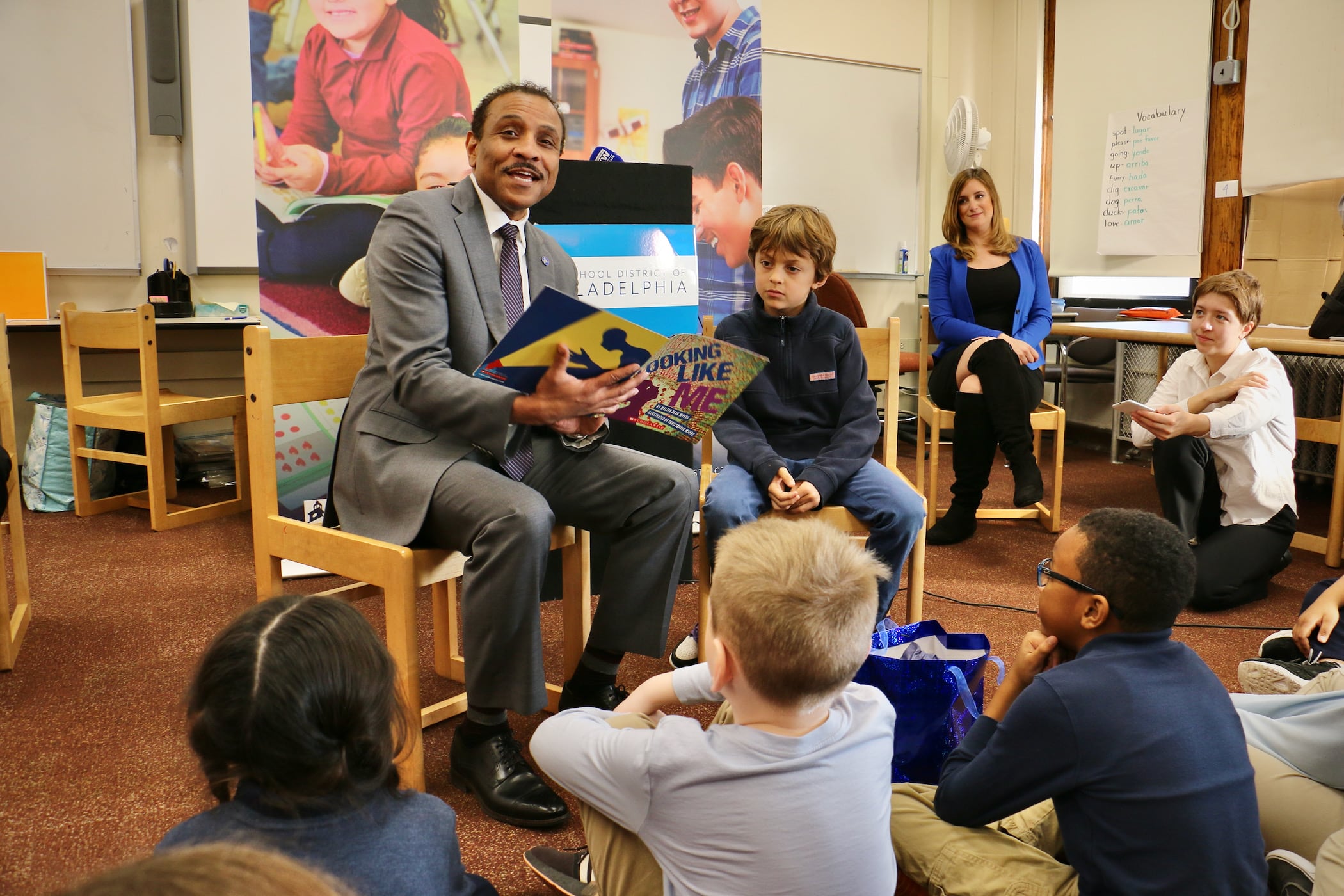 A man in a gray suit and dark tie sits on a chair reading a book aloud. A boy in a blue shirt and jeans sits next to him. Students sit on the floor listening. A teacher wearing a black dress, black boots, and a bright blue jacket sits to the left of him.