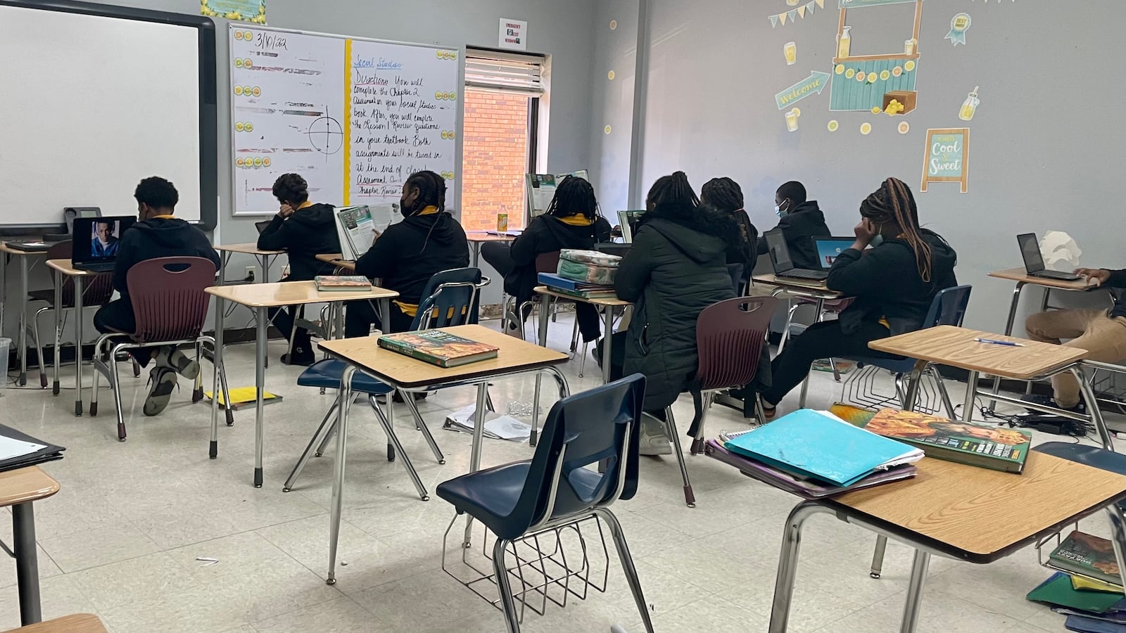 A back view of students working at their desks, facing a smart board at the front of a a classroom.