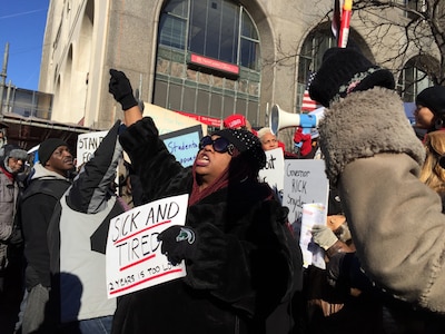 A photograph of adults wearing jackets and holding protest signs outside.