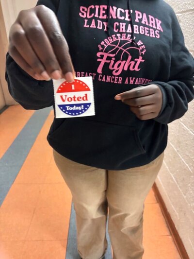 A young female student wearing a black hoodie with pink letters holds up an "I Voted Today" sticker.