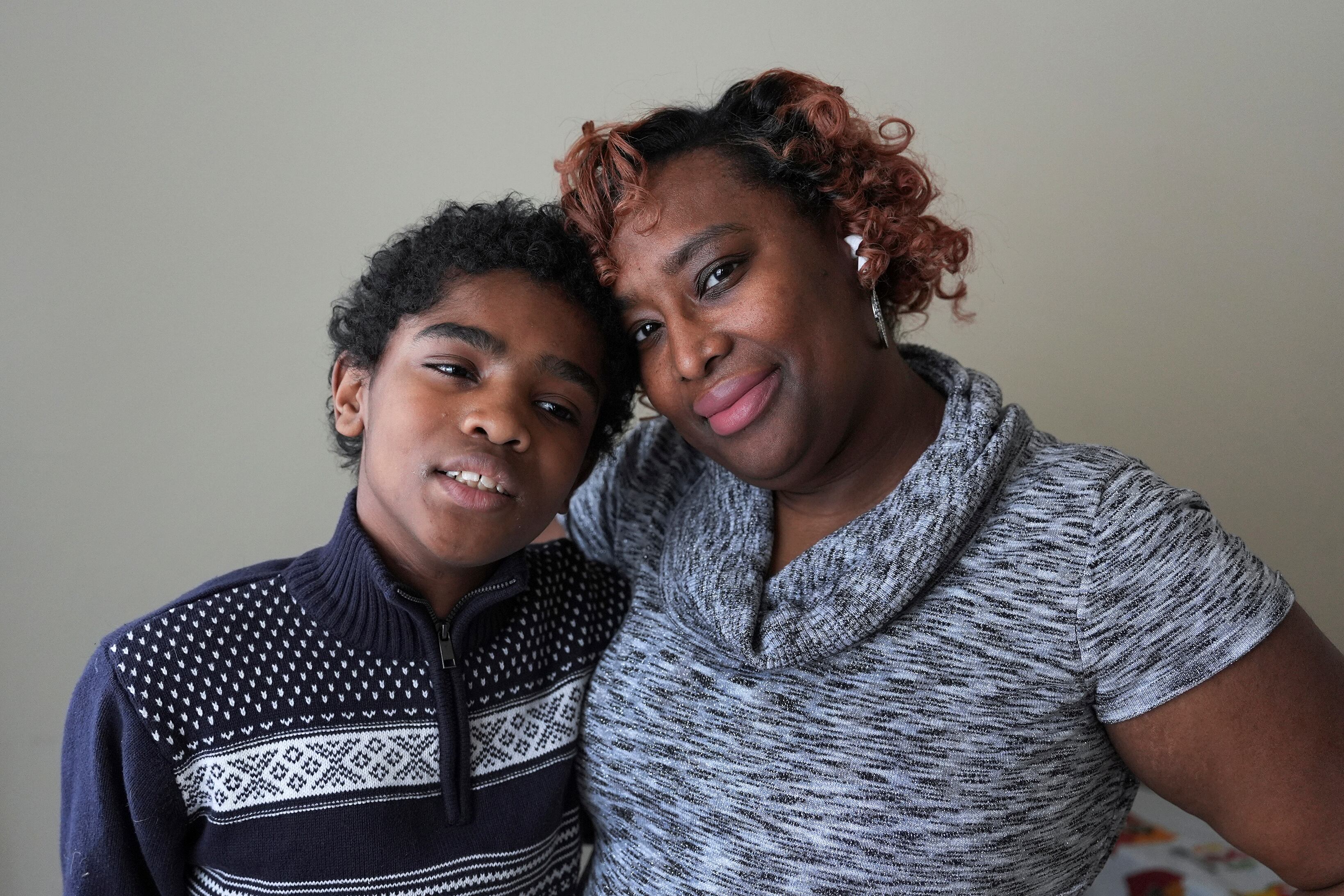 A mother with short curly hair and a preteen son, embrace for a portrait against a grey wall.