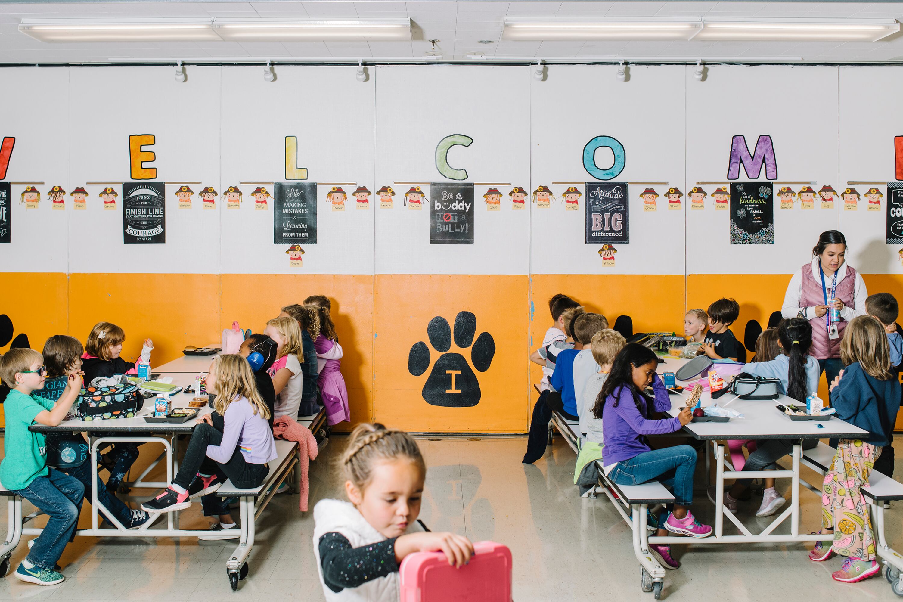 A sign that says "Welcome" in colorful letters on the back white and orange wall of a school cafeteria with two large tables filled with young elementary students eating lunch.