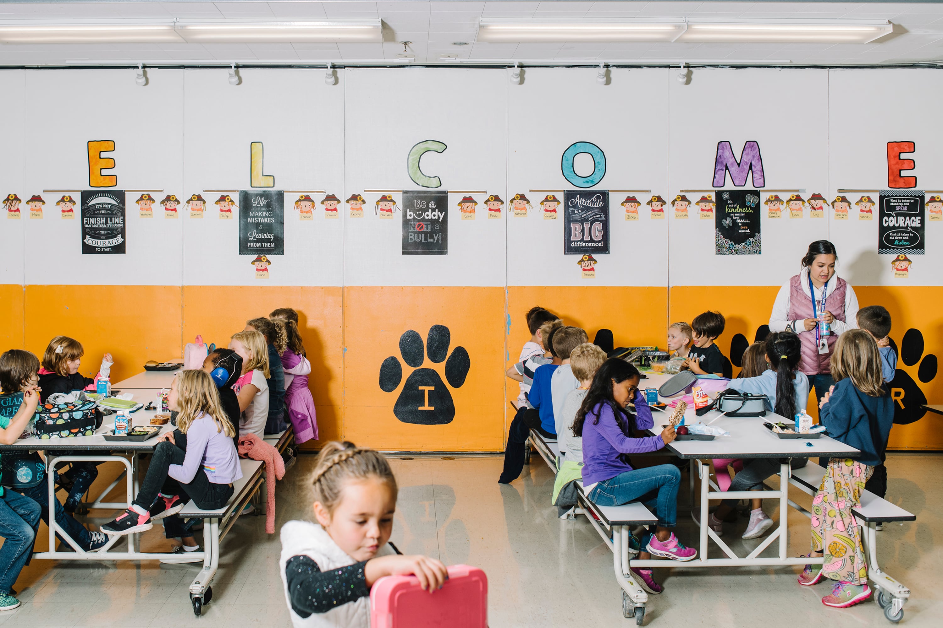 A sign that says "Welcome" in colorful letters on the back white and orange wall of a school cafeteria with two large tables filled with young elementary students eating lunch.