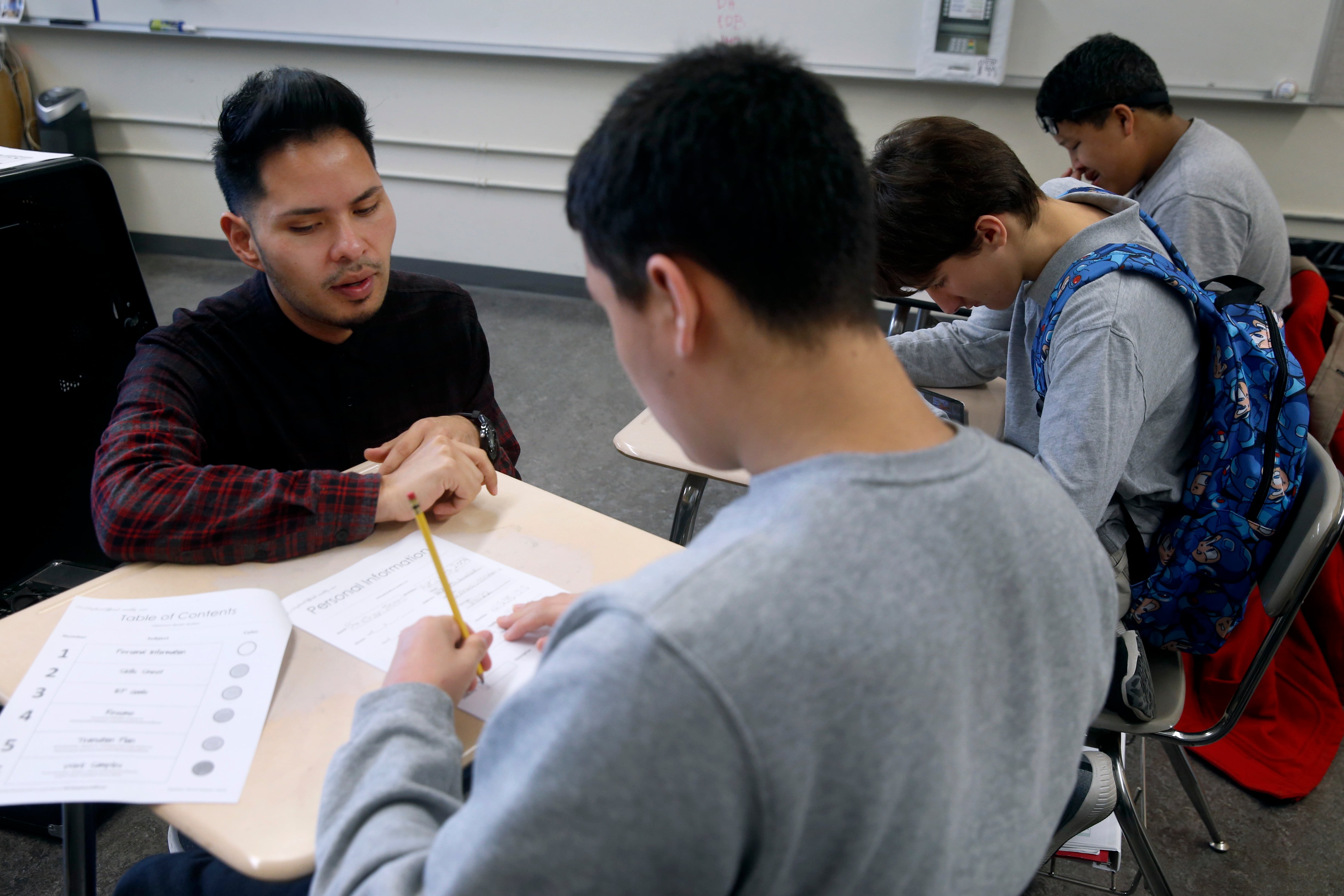 A man in a dark shirt watches as a student in a gray shirt writes on a paper.