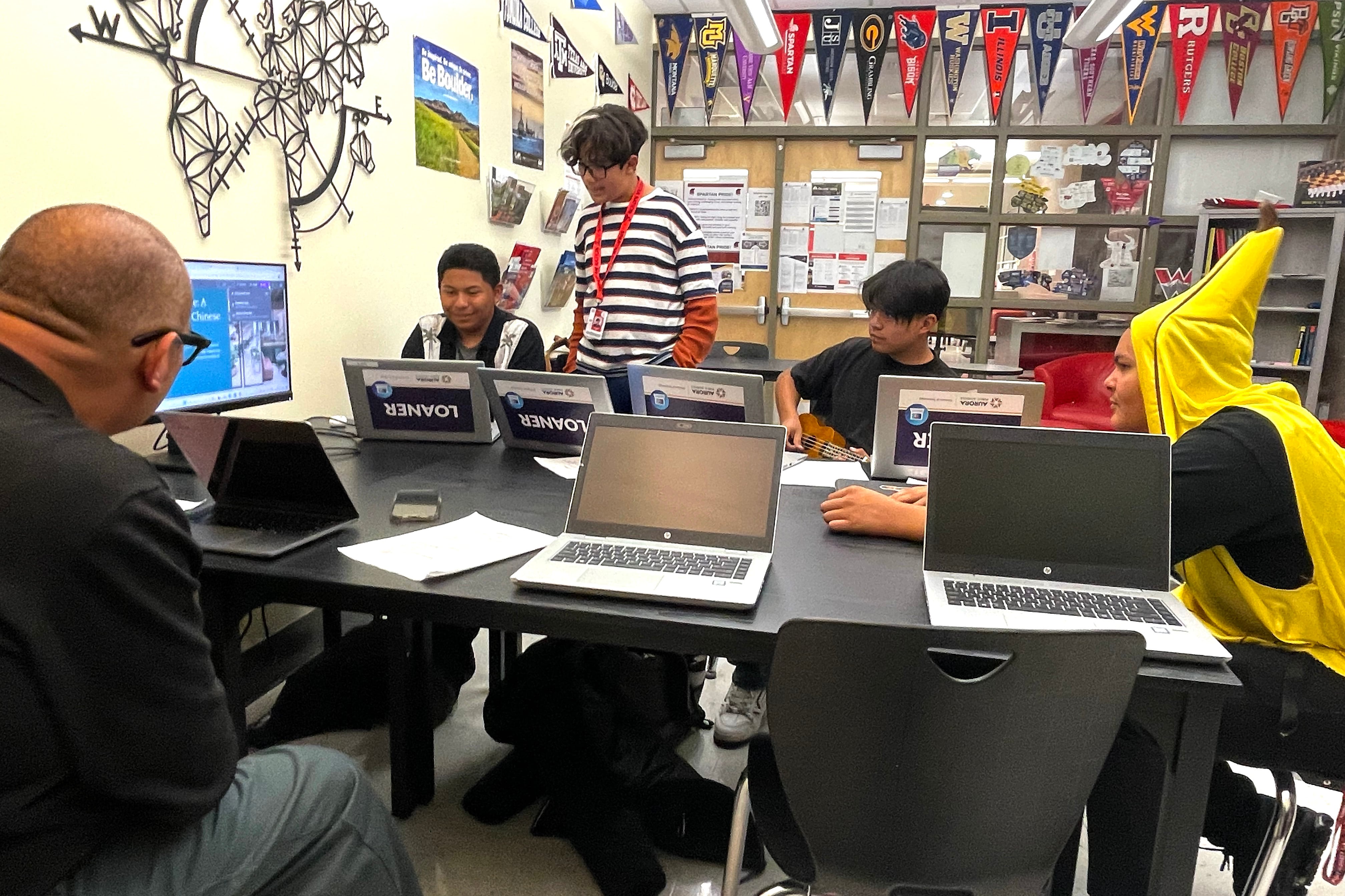 Students -- one dressed for Halloween in a banana costume -- and a man sit at tables in a classroom setting.