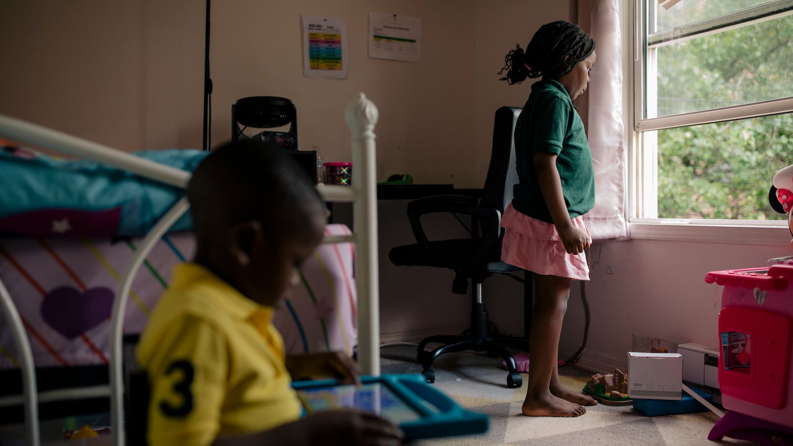 A young girl, wearing a green shirt and pink skirt, looks out her bedroom window as her brother sits on the floor next to her bed.