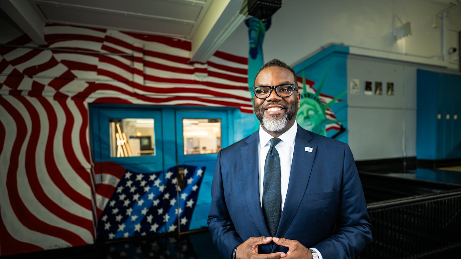 Chicago Mayor Brandon Johnson smiles while standing in a school hallway with a mural of an American flag behind him.
