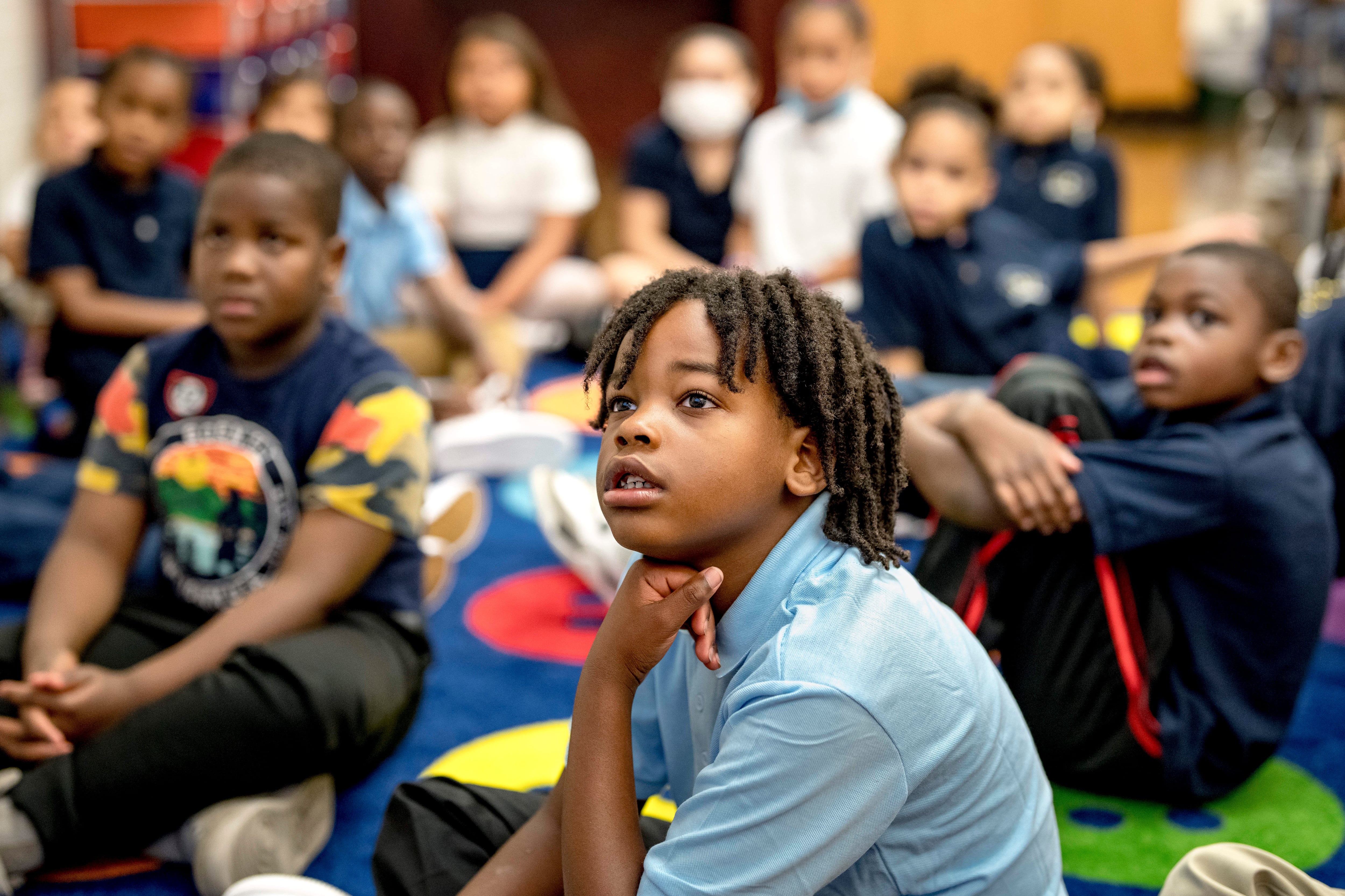 A student looks up toward a video while sitting on the floor, as other students sit behind him.