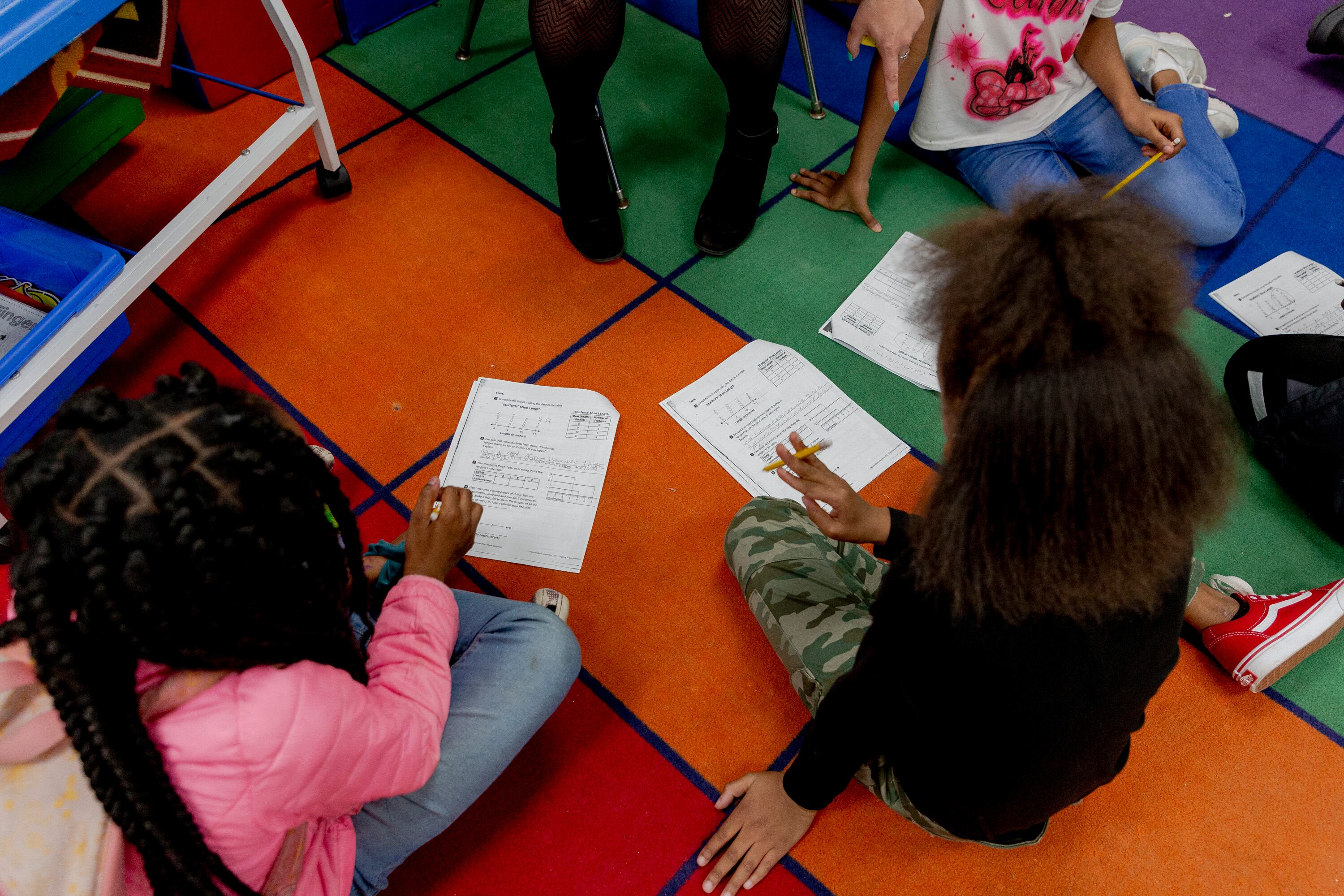 Two young girls work on math worksheets while sitting on a brightly colored rug with the legs of their tutor in black stockings visible nearby.