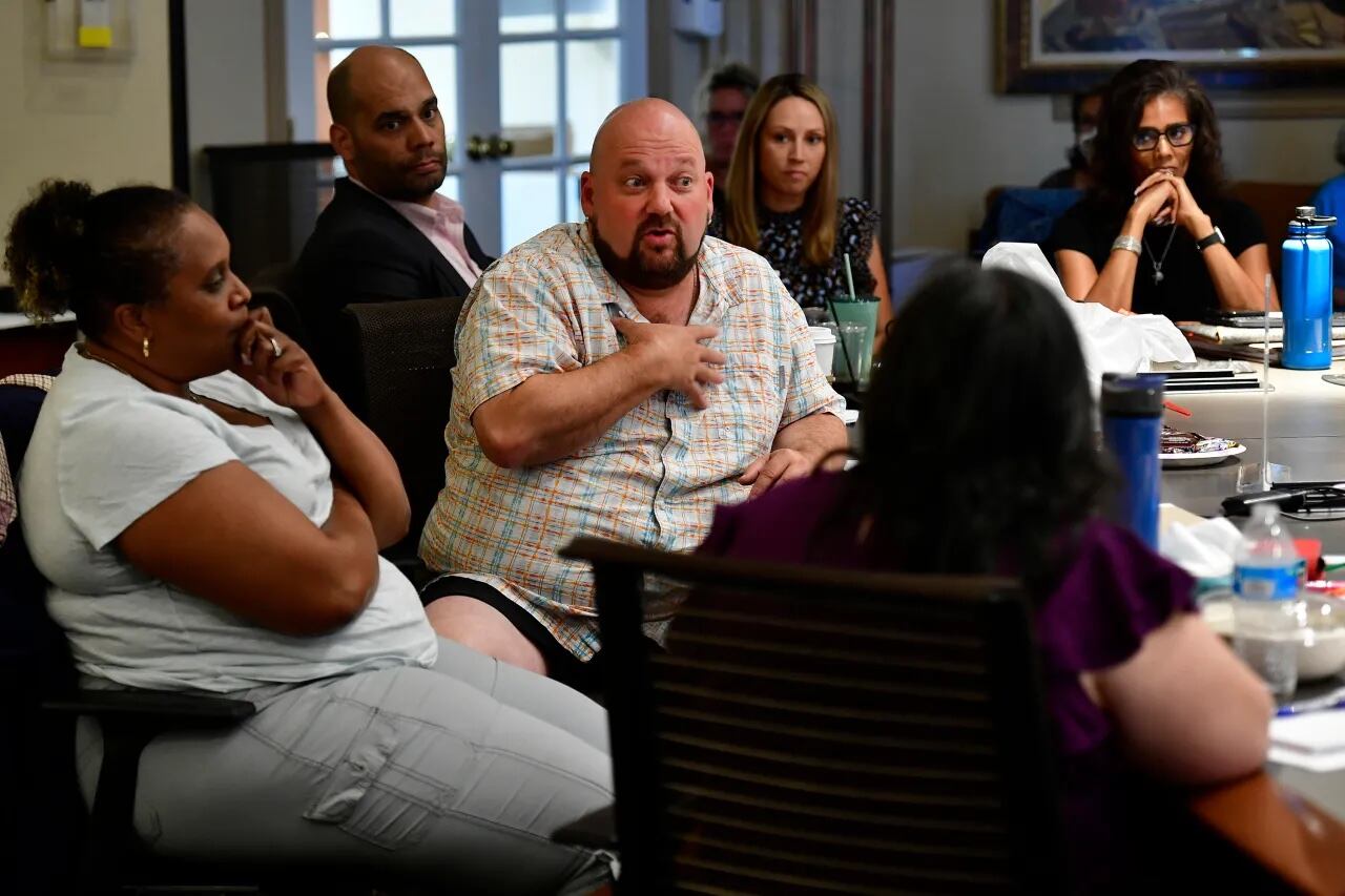A man wearing a button-down shirt gestures to himself while speaking during a school board meeting. His eyes are wide.