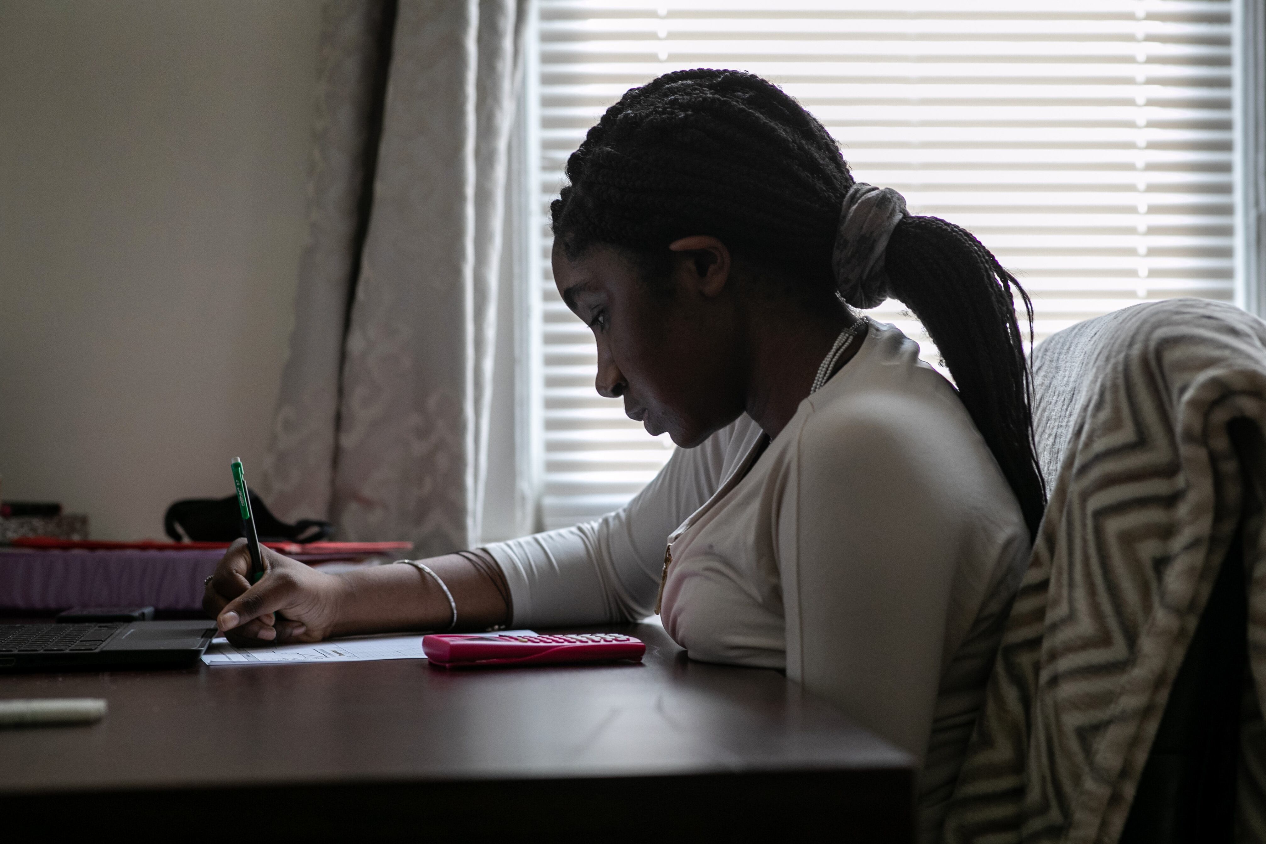 A teenage girl with braided hair takes notes while virtually learning at her desk.