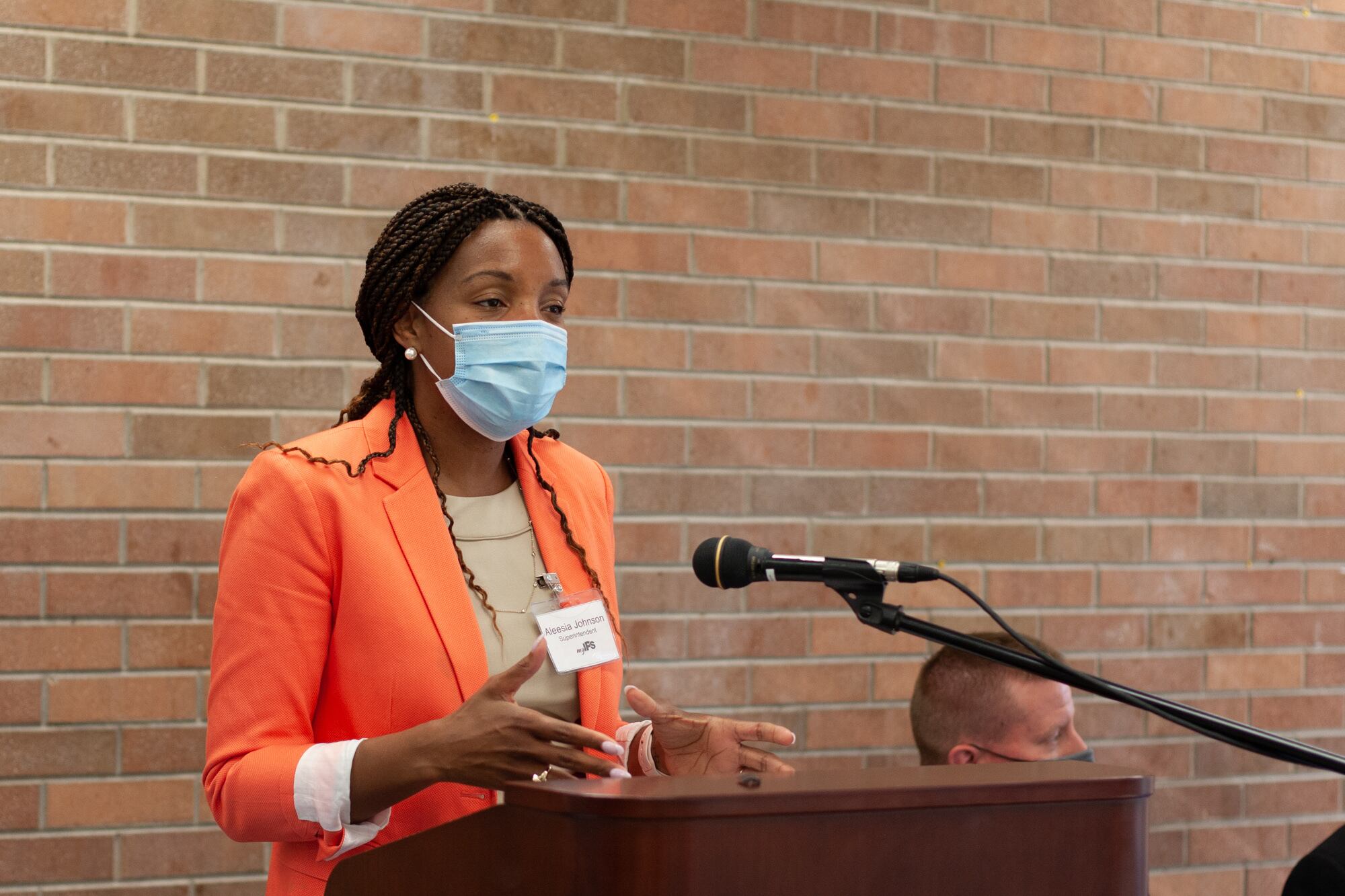 A woman in a formal bright orange jacket and a beige shirt wearing a blue face mask gestures with her hands as she speaks into a microphone at a podium in front of a red brick wall