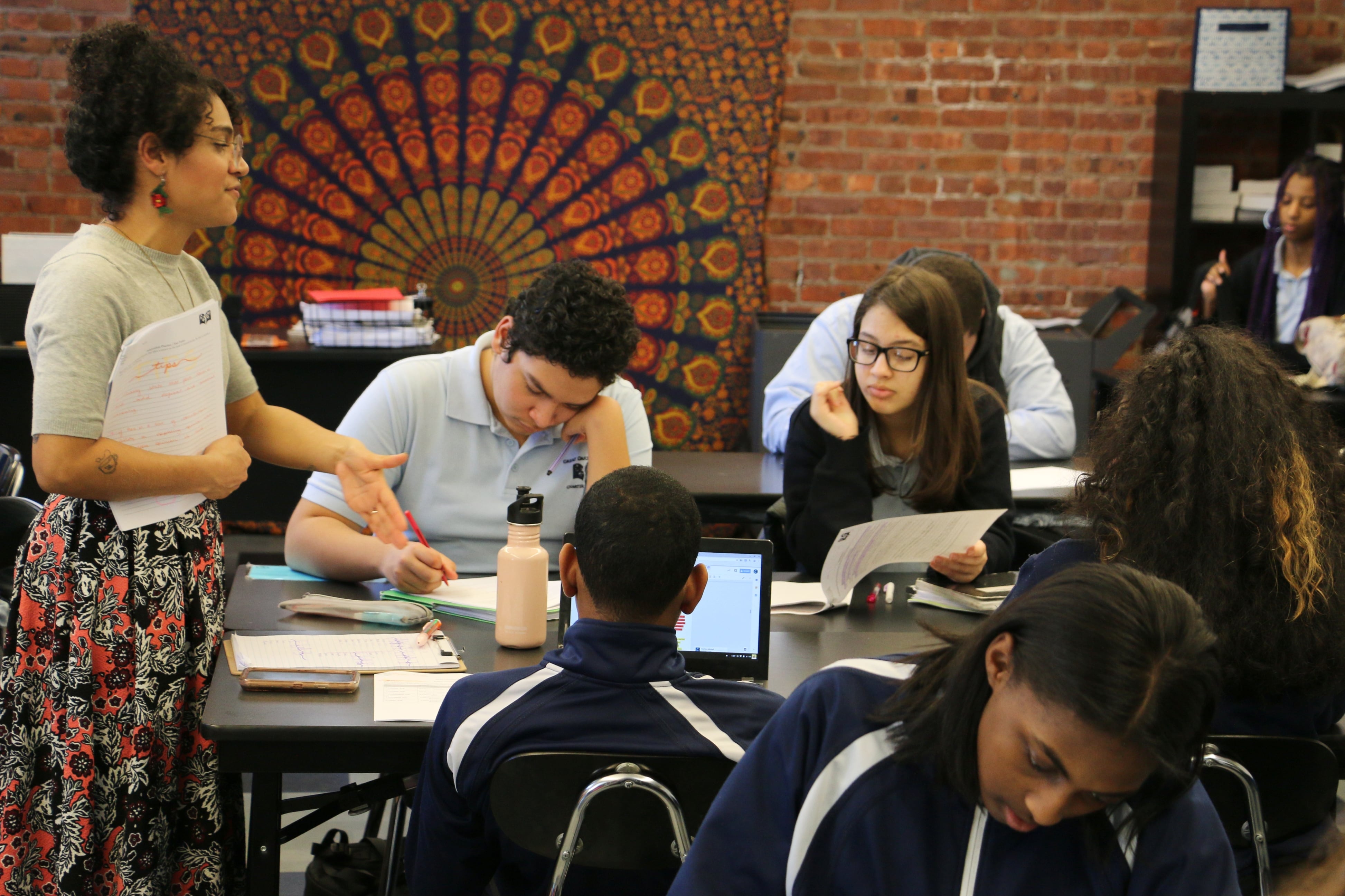 A woman wearing a flowery skirt speaks to a group of four students who are seated at tables and writing.