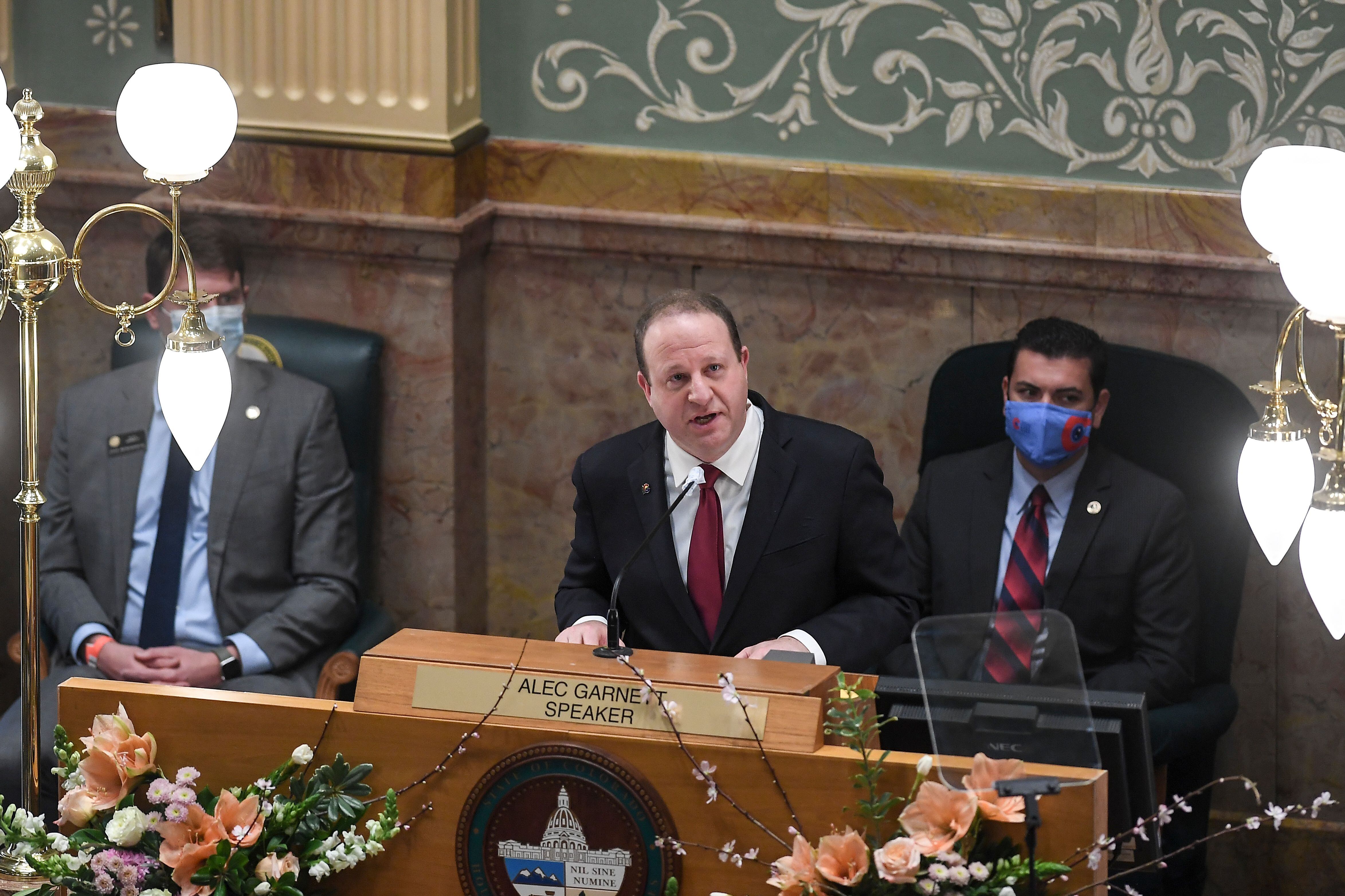 Gov. Jared Polis, flanked by two masked officials, stands at a lectern speaking.