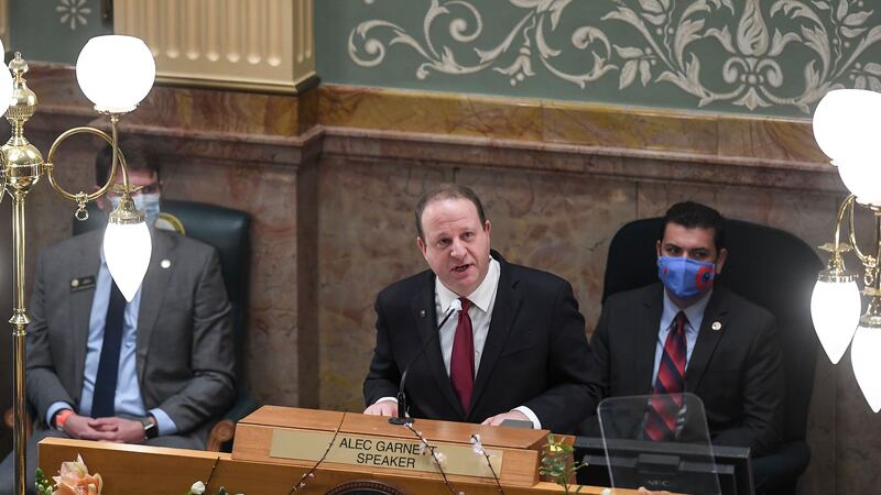 Gov. Jared Polis, flanked by two masked officials, stands at a lectern speaking.