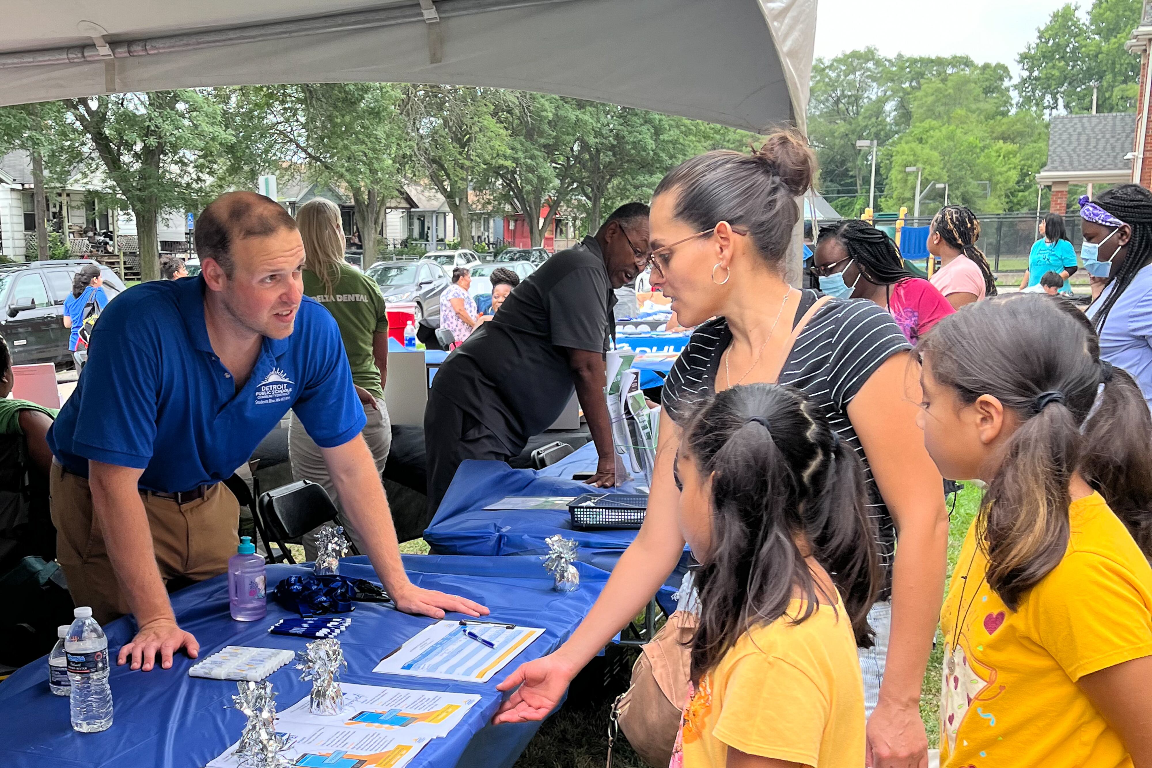 A man working for Detroit Public Schools Community District stands at a table distributing papers to a mother and her two daughters.