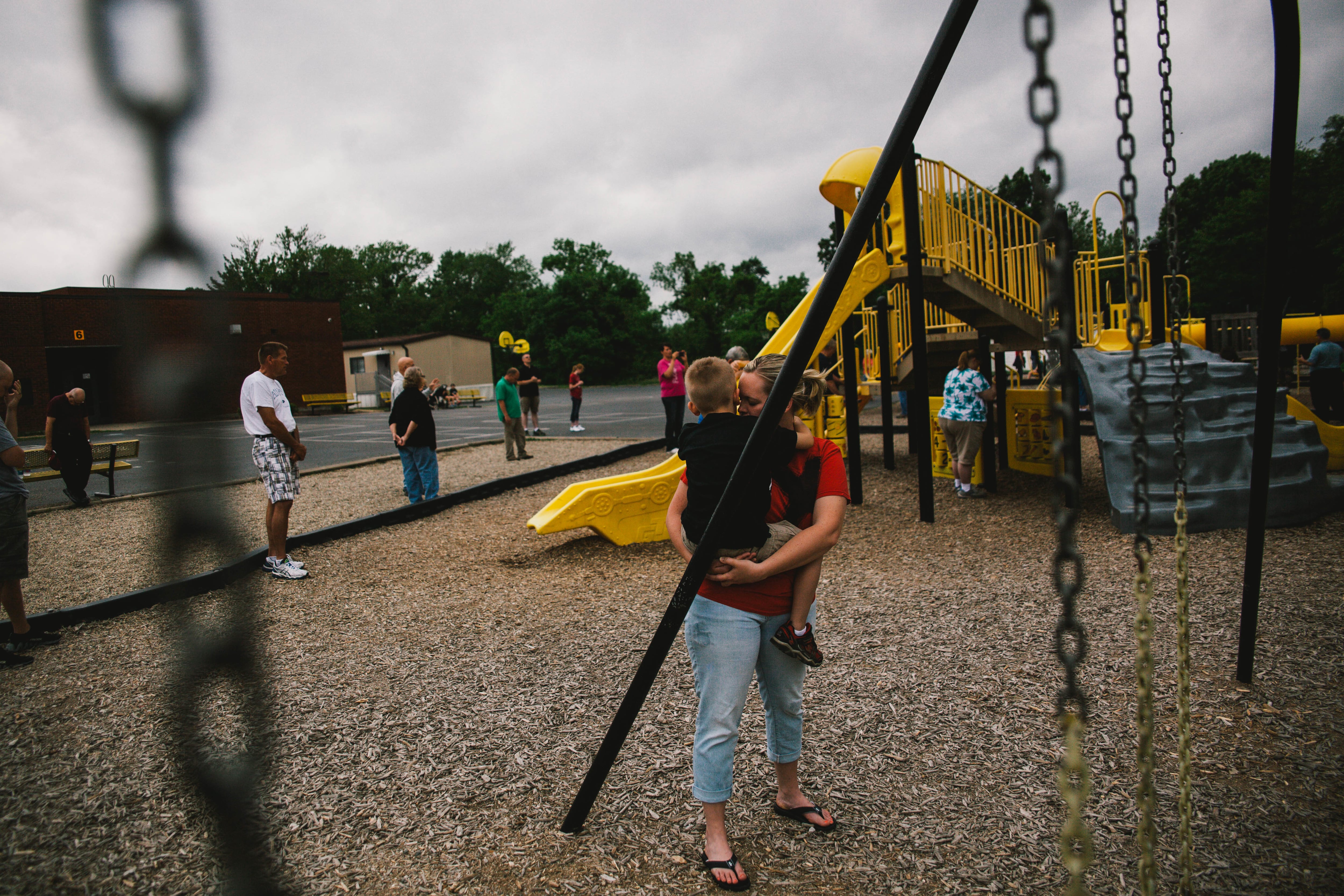 A woman holds her child next to a set of swings, as a circle of people pray around the edge of the playground.