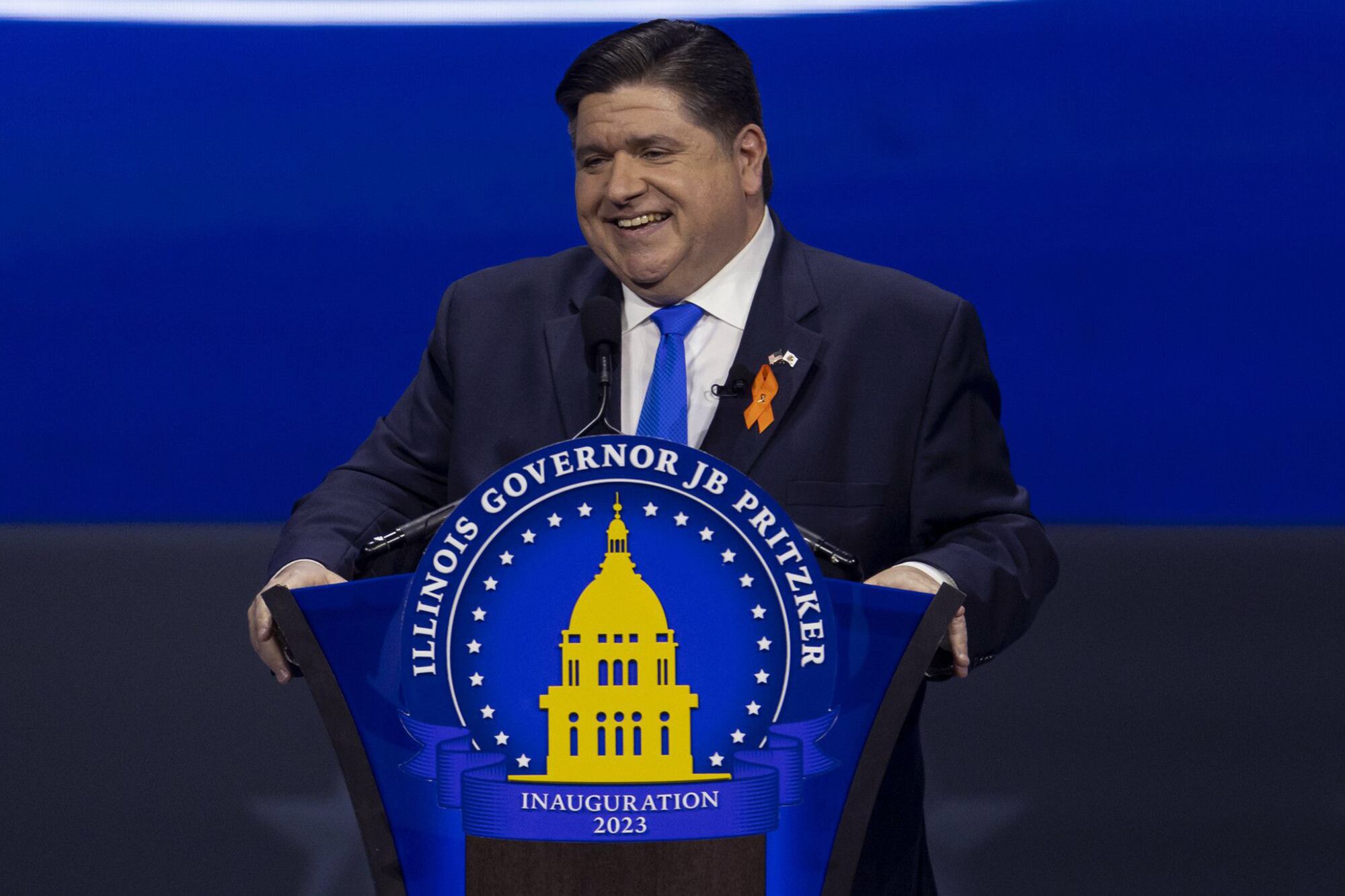 A man with short dark hair stands at a podium with an emblem in the front and a blue background.
