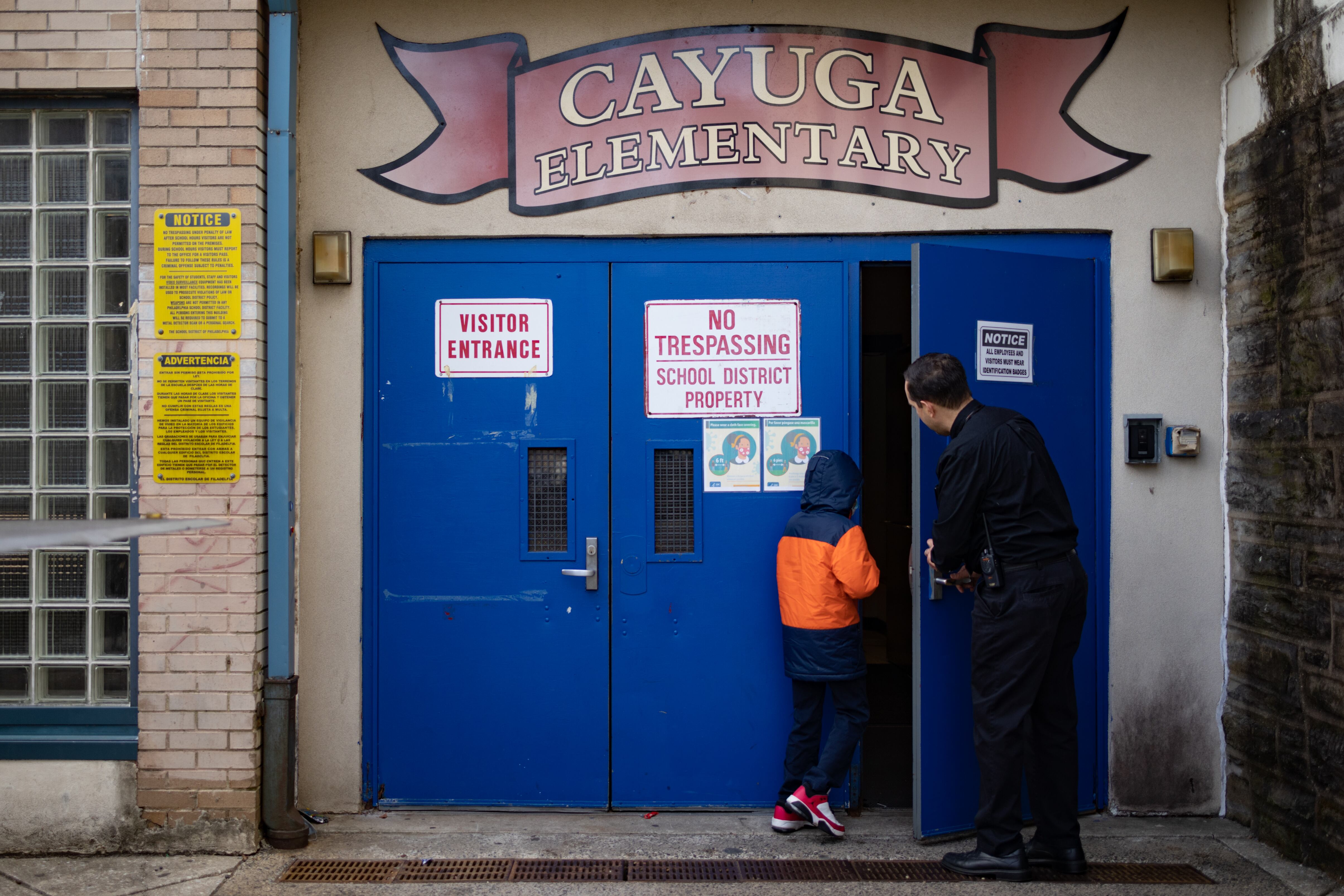 A man in dark clothing holds a blue door open as a child in an orange and blue coat and red sneakers enters a building with a “Cayuga Elementary” sign above the door.