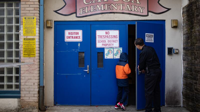A man in dark clothing holds a blue door open as a child in an orange and blue coat and red sneakers enters a building with a “Cayuga Elementary” sign above the door.