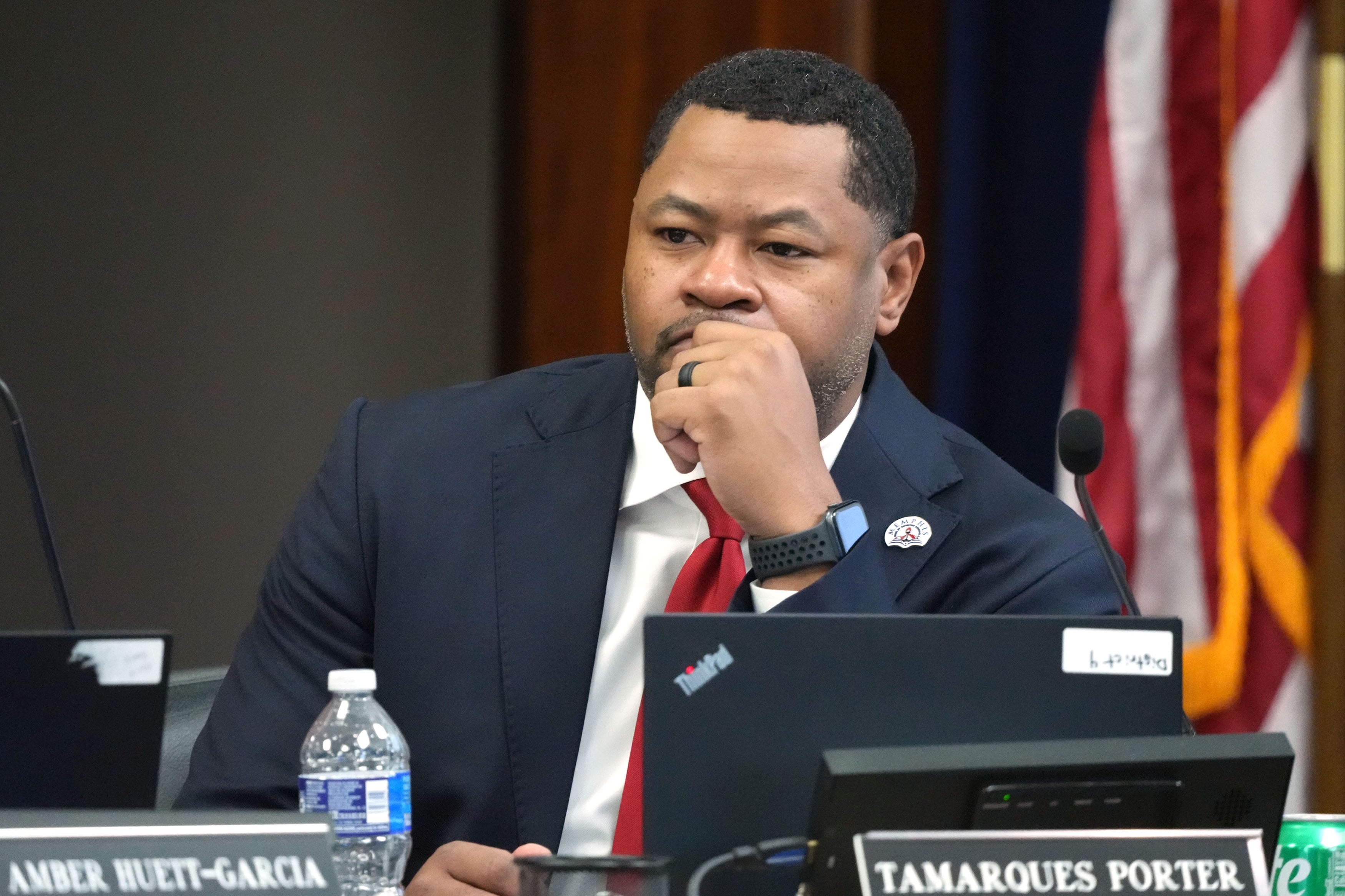 A photograph of a Black man in a suit sitting at a wooden desk in a large conference room.