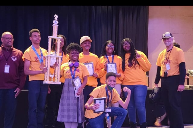 Students stand on a stage and hold a trophy they just won at the national chess championships.