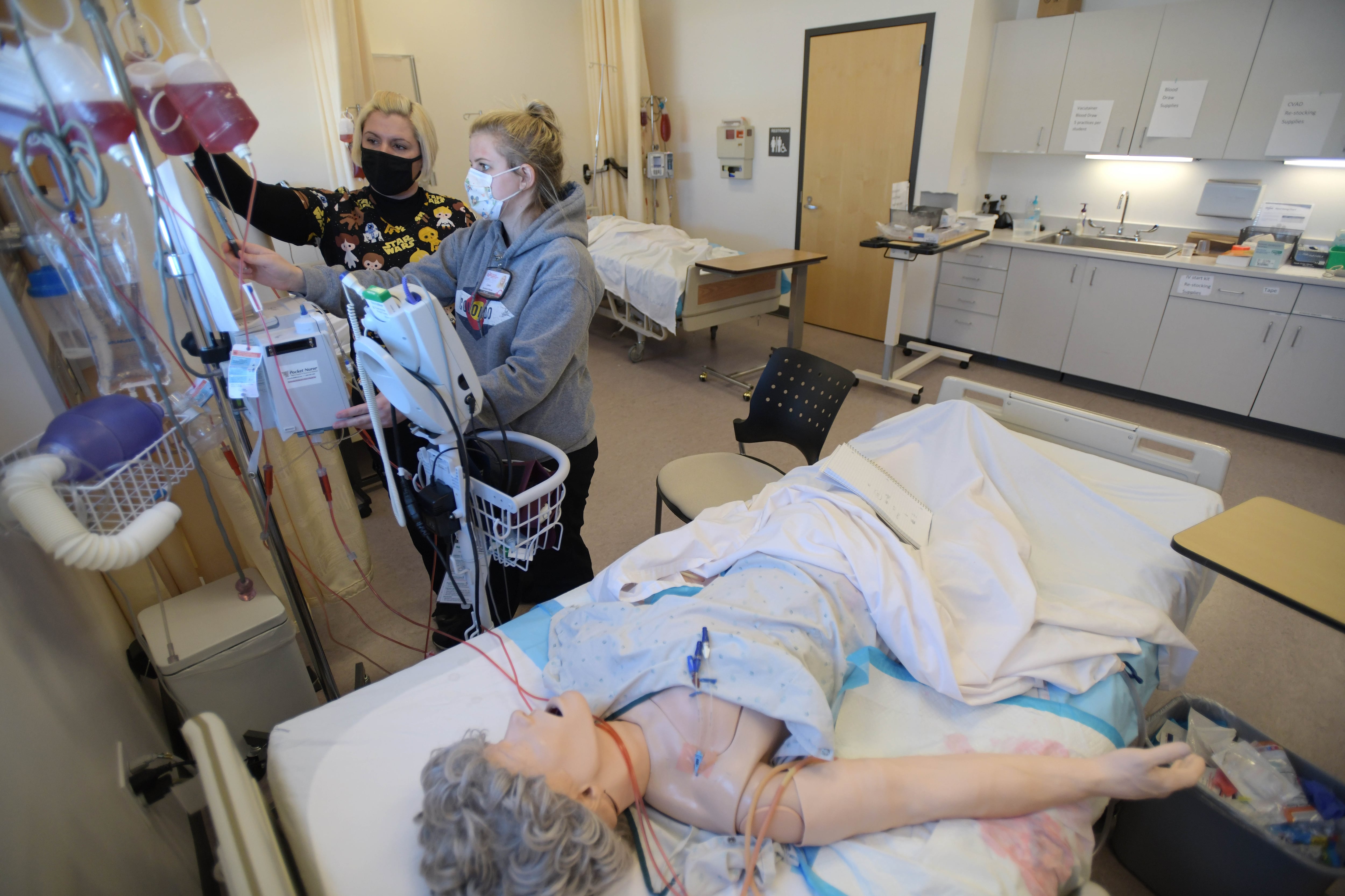 Two masked nursing students, Jade Prophet, left, and Cami Gardetto, stand at a monitor in a nursing simulation classroom at Colorado Northwestern Community College. In the foreground a dummy of a woman lays on a hospital bed, with tubes connecting her to machines.