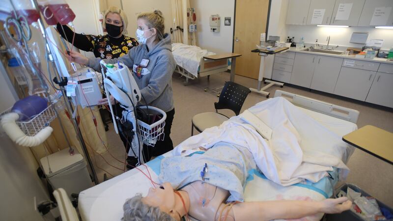 Two masked nursing students, Jade Prophet, left, and Cami Gardetto, stand at a monitor in a nursing simulation classroom at Colorado Northwestern Community College. In the foreground a dummy of a woman lays on a hospital bed, with tubes connecting her to machines.