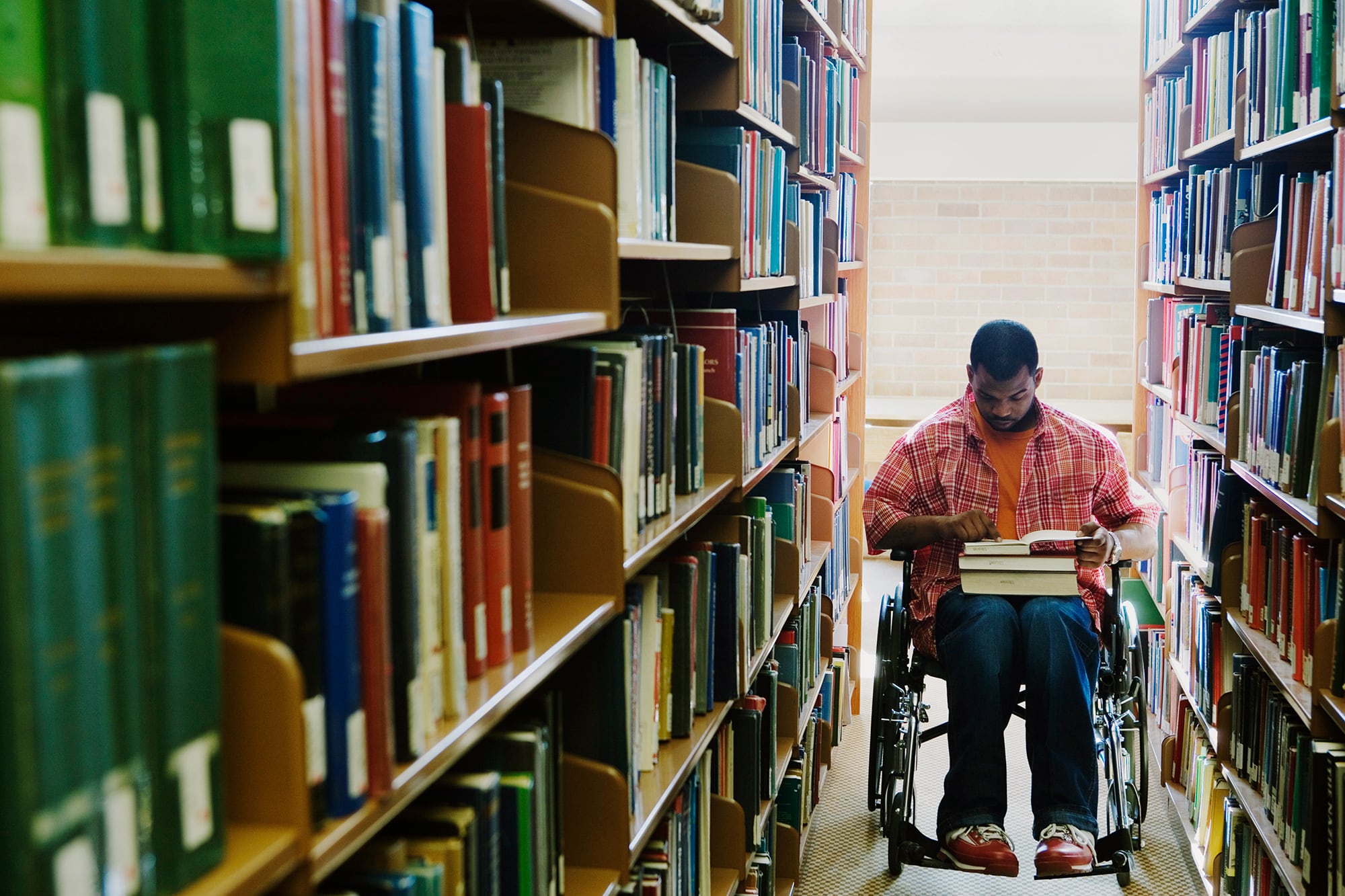 A man in a wheelchair looks at a book in his lap while in between two shelves filled with books. 