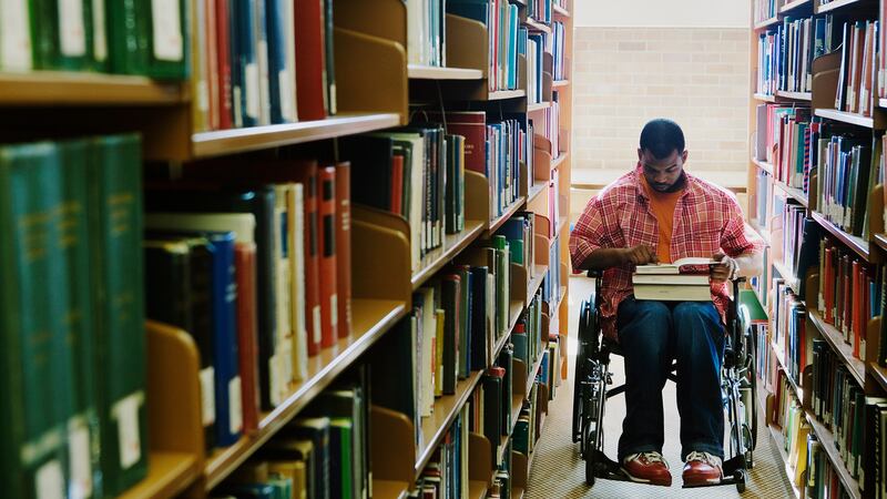 A man in a wheelchair looks at a book in his lap while in between two shelves filled with books. 