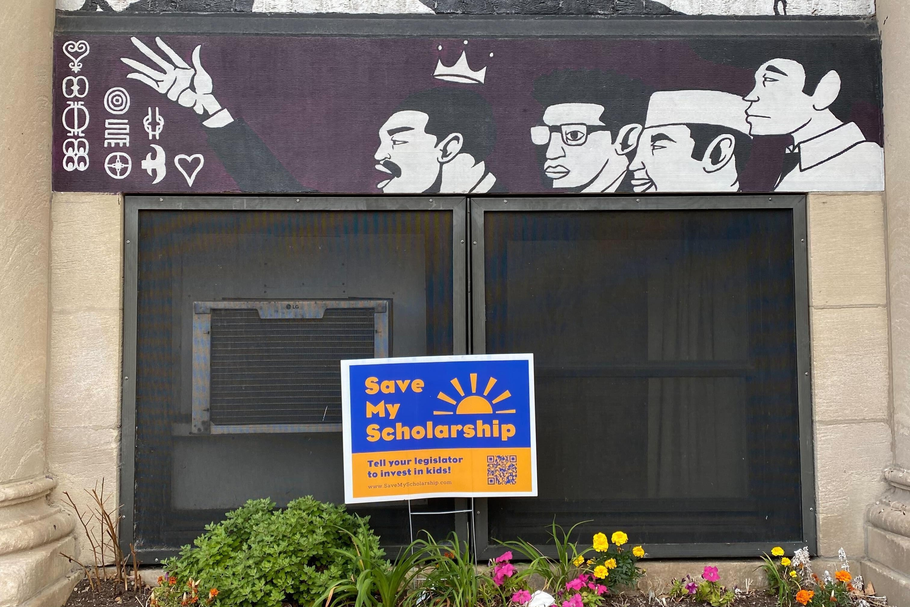 A blue and yellow sign that reads "save my scholarship" sits in a flower bed with a mural in the background on a school building.