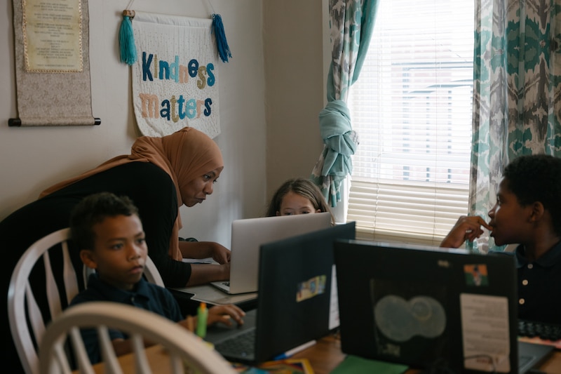 A woman in a hijab stands behind two young children on computers