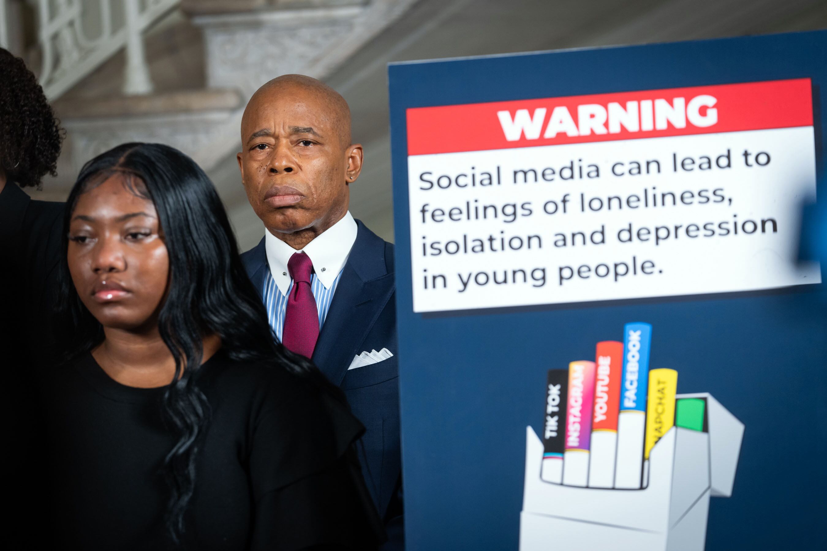 A man in a suit and tie stands next to a sign with health warnings about social media.