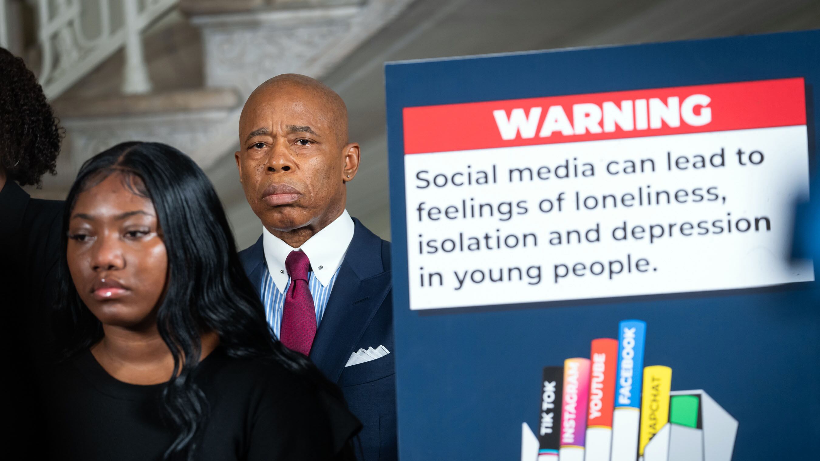 A man in a suit and tie stands next to a sign with health warnings about social media.