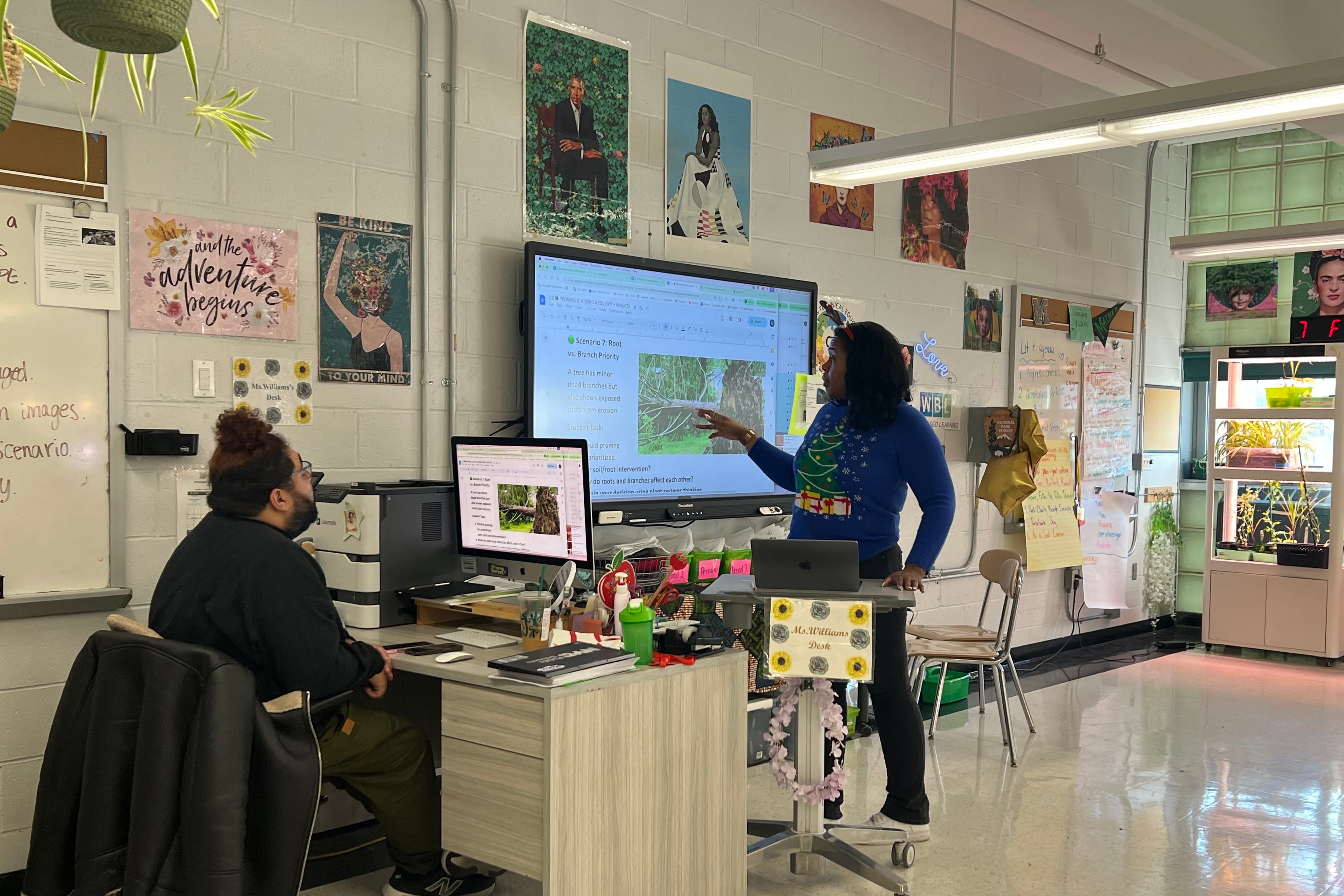 A man with dark hair sits at a desk working on a computer. To his right a woman is standing up teaching a class.