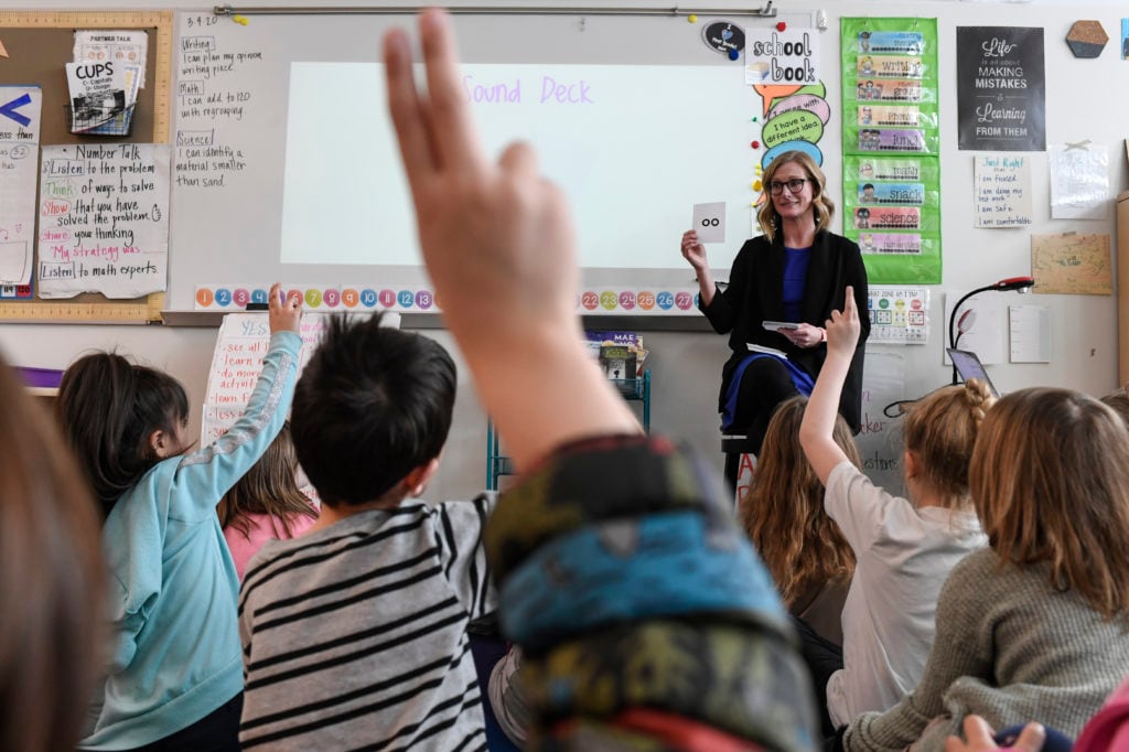 A first-grade teacher faces her students, who are seated on the rug with their backs to the camera.
