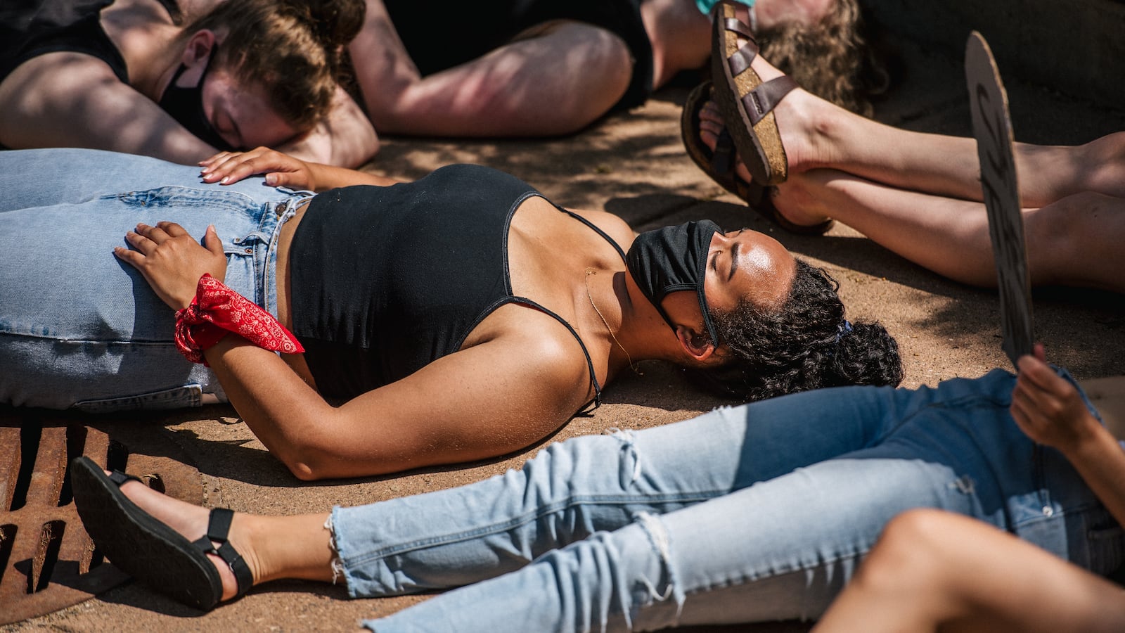 A group of young students lie on the ground during a die-in protest. The young woman in the center is wearing blue jeans, a black tank top and a red scarf on her arm. Other students’ limbs are all around her.
