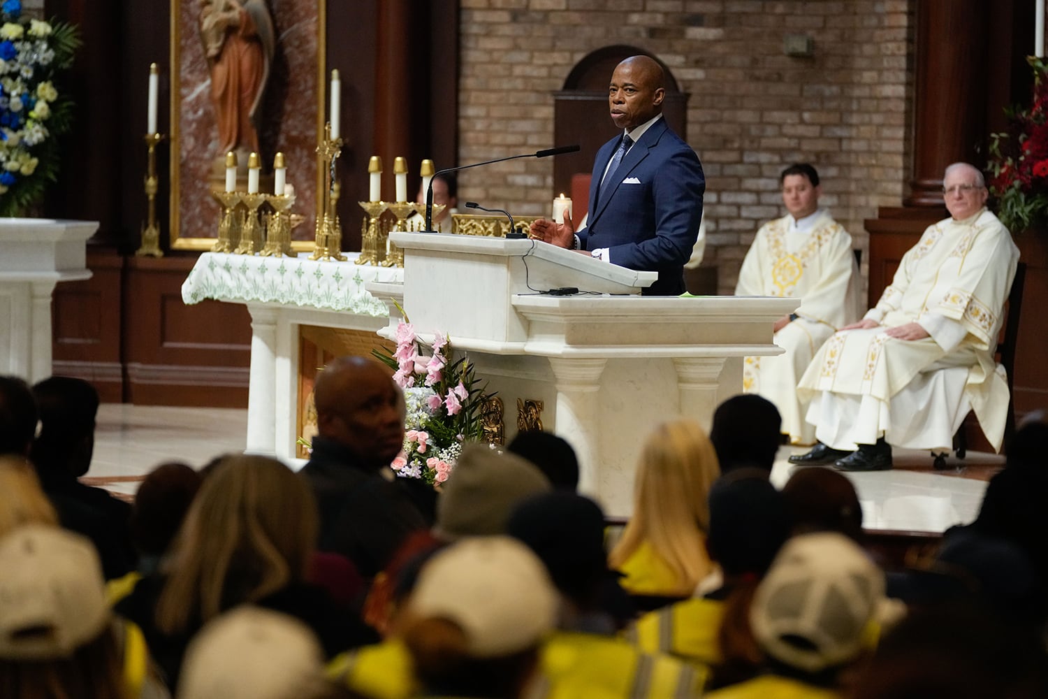 A man wearing a blue suit stands at a podium inside of a church. There are two church officials wearing all white in the background and people sitting in pews in the foreground.