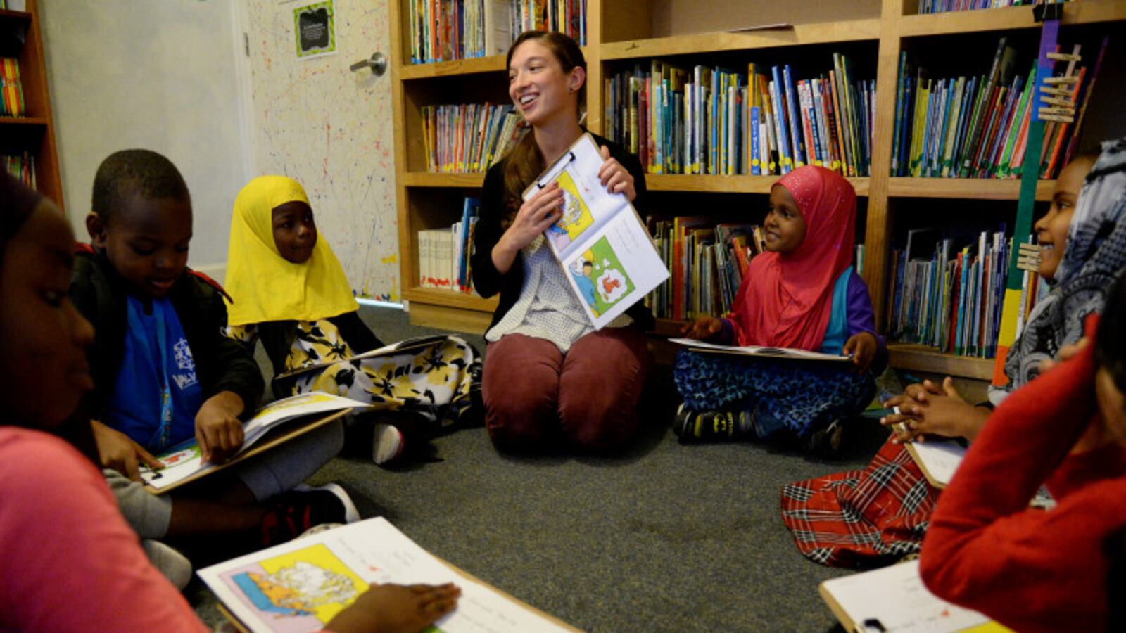 A young woman smiles as she shows a picture book to a group of young children seated in a circle beside her.