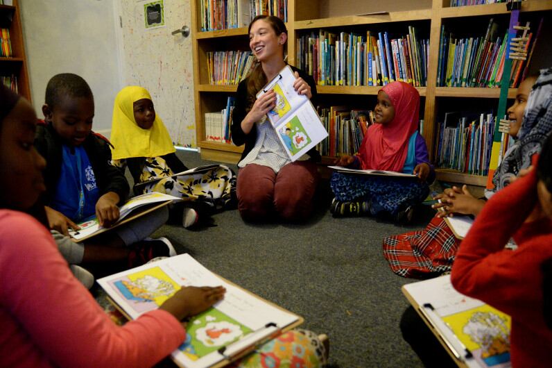 A young woman smiles as she shows a picture book to a group of young children seated in a circle beside her.