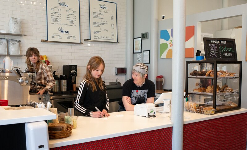 A 12-year-old girl with long straight hair writes on a piece of paper on the counter of a coffee shop. She’s behind the counter with two adult women. One of the women, with short, light hair and a snazzy crown, leans on the counter talking with the girl.