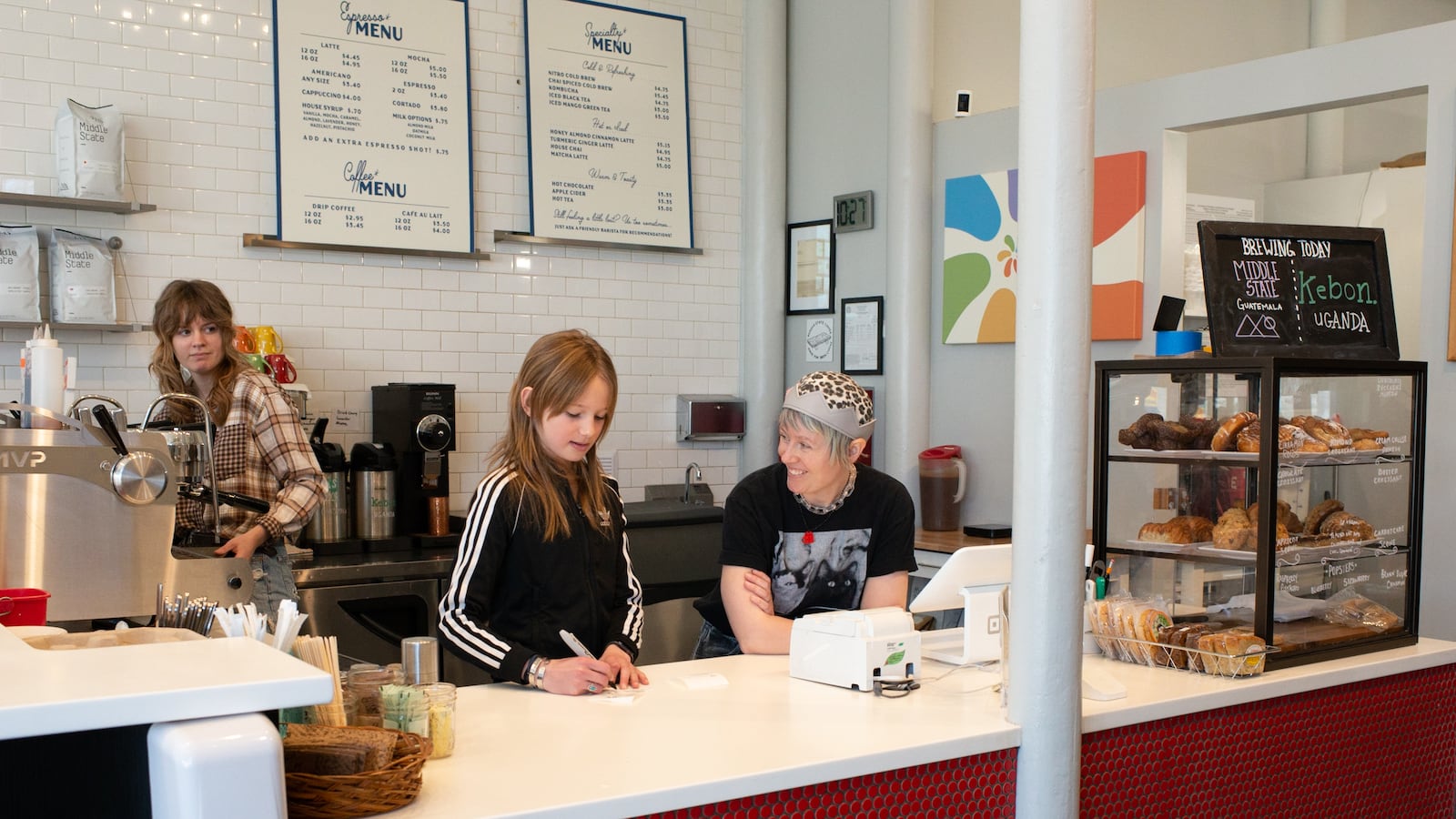 A 12-year-old girl with long straight hair writes on a piece of paper on the counter of a coffee shop. She’s behind the counter with two adult women. One of the women, with short, light hair and a snazzy crown, leans on the counter talking with the girl.