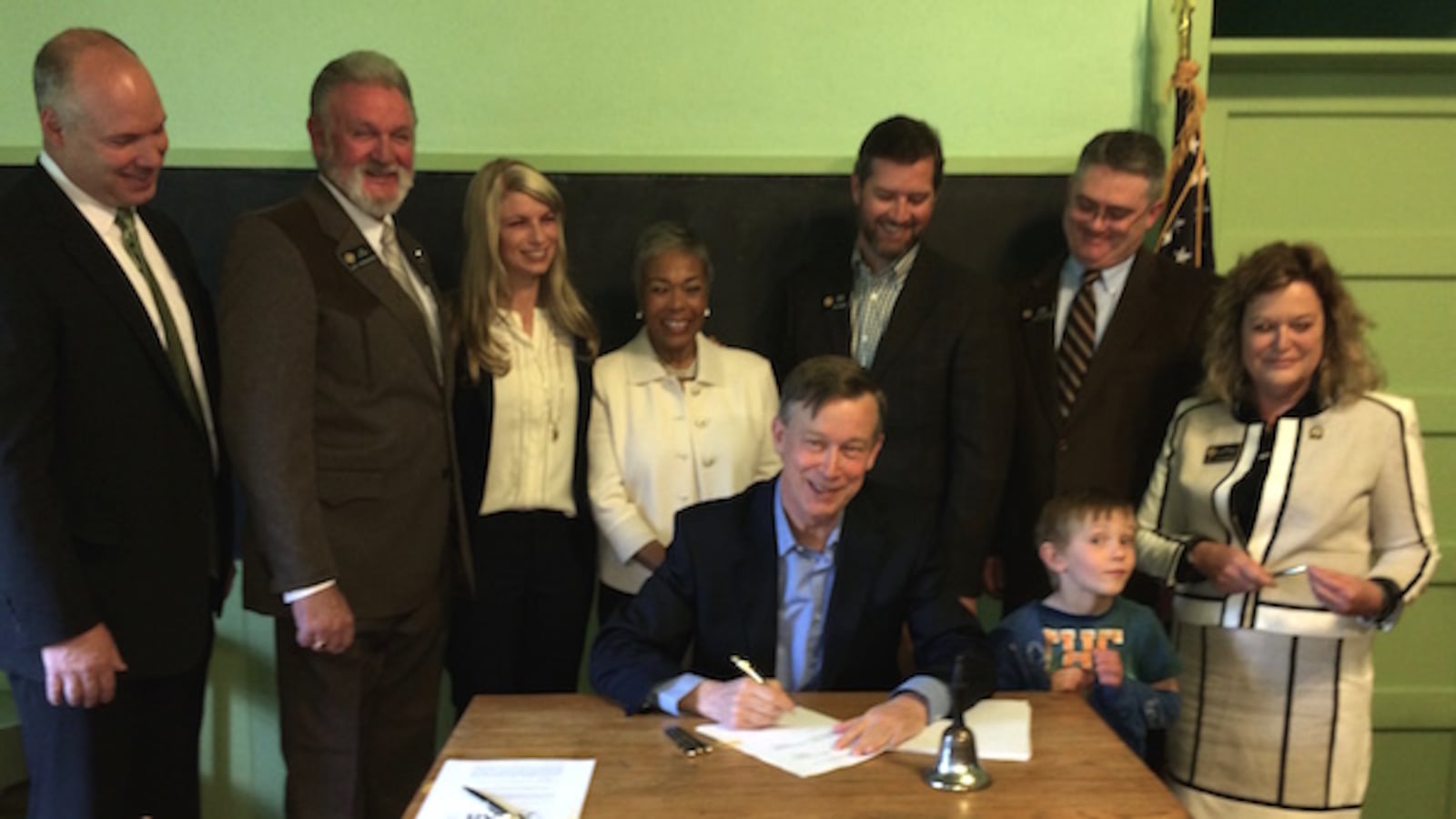 Gov. John Hickenlooper signs a test bill flanked (L-R) by Reps. Kevin Priola, Jim Wilson, Brittany Pettersen, Janet Buckner (wife of Rep. John Buckner), Sens. Andy Kerr and Chris Holbert and Rep. Tracy Kraft-Tharp. Kerr's son Griffin mugs for the cameras.