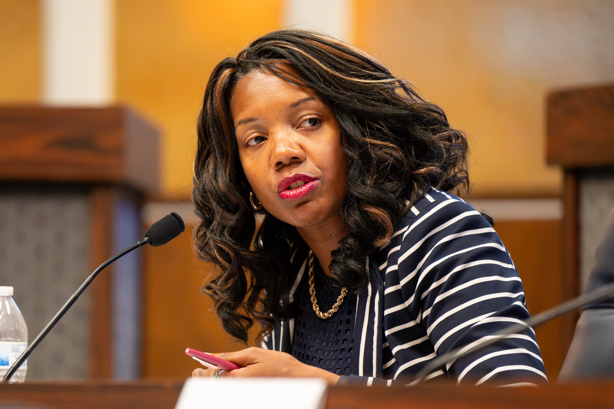 A photograph of a Black woman wearing a black and white striped sweater speaking into a microphone while sitting at a wooden desk in a large meeting room.