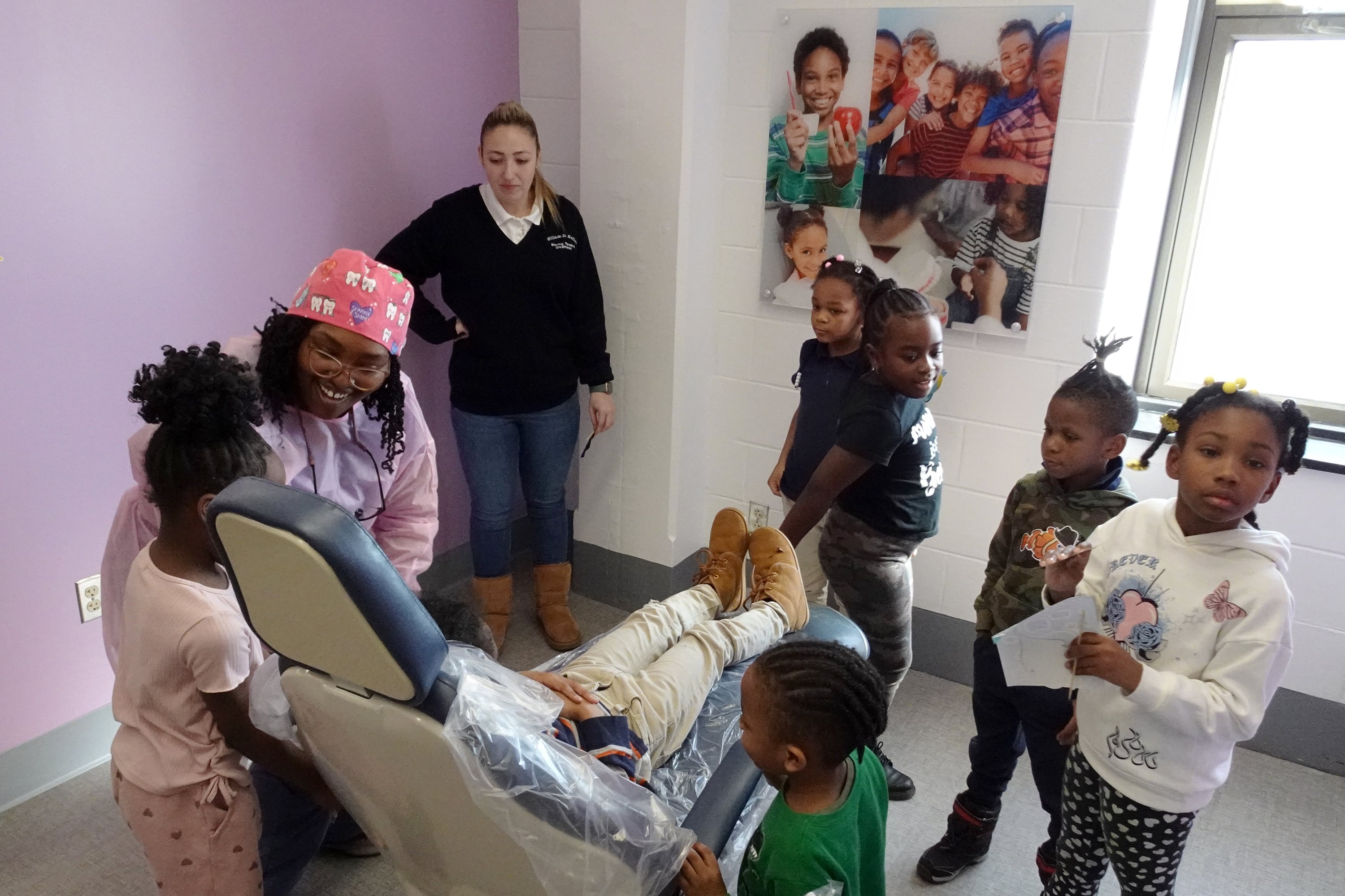 A young child sits in a dentist chair while and adult wearing scrubs and a group of young children stand around.
