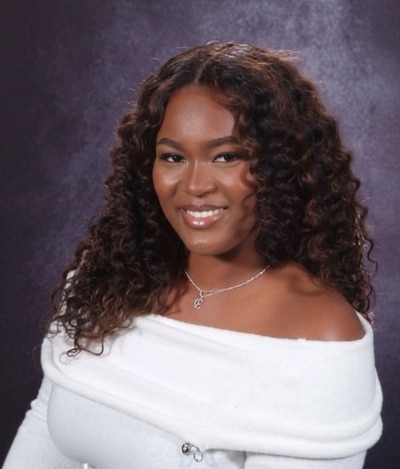 A photograph of a Black high school student with long, dark, curly hair and wearing a white sweater while smiling and posing for a portrait against a purple backdrop.