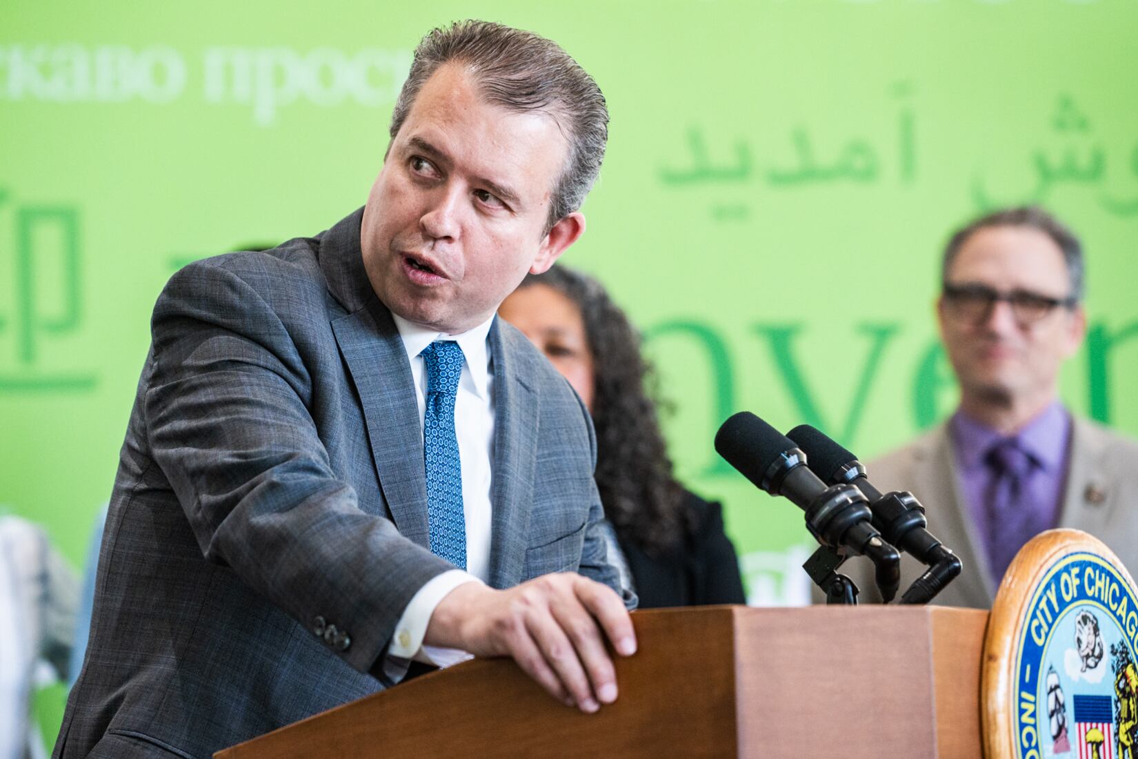 A man wearing a suit speaks from behind a wooden podium with a microphone attached to the front.